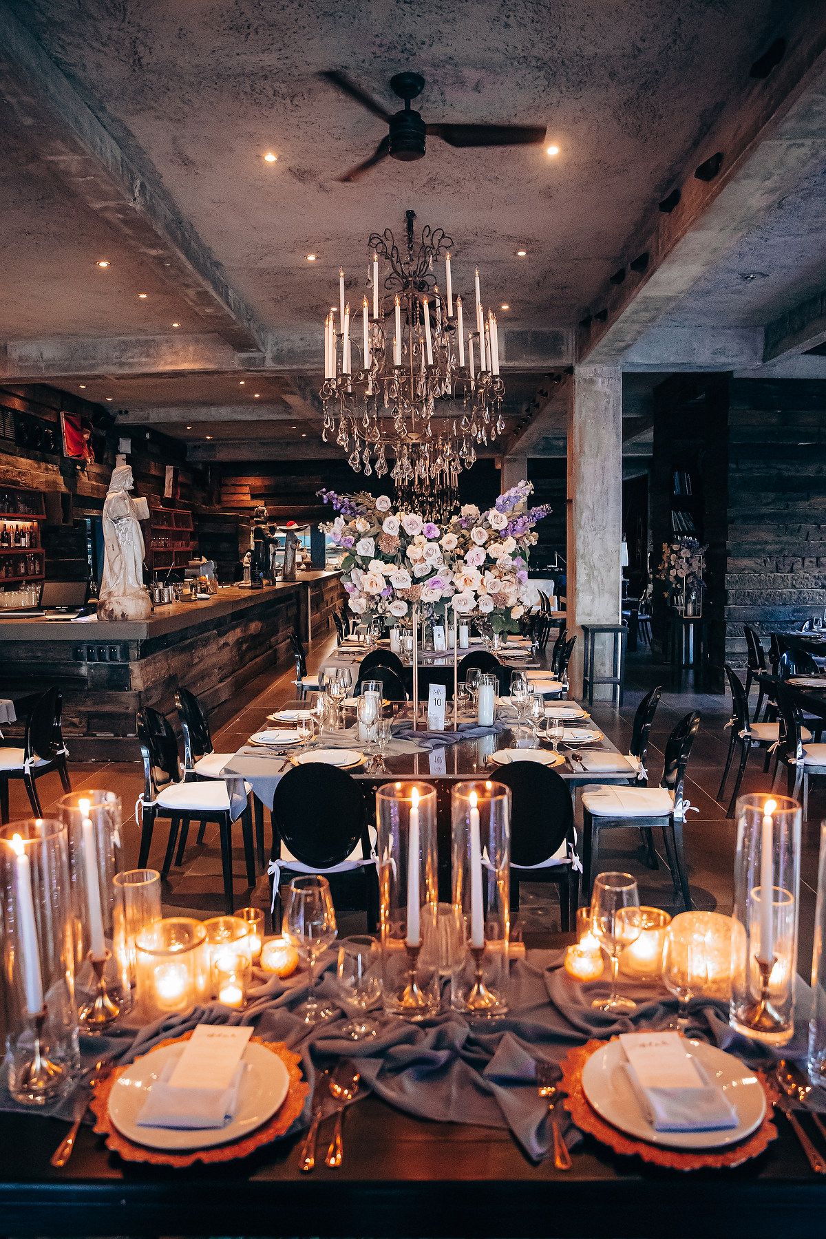 Elegant candlelit dining tables set with white linens and crystal chandeliers in a rustic industrial restaurant space.