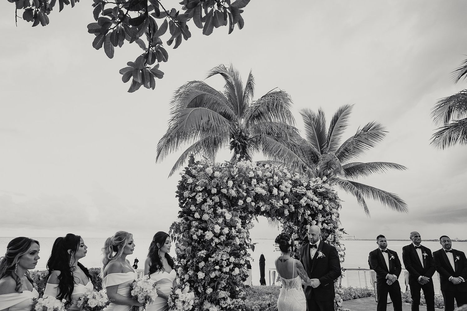 Black and white tropical beach wedding ceremony with palm trees and guests gathered on sandy shore.