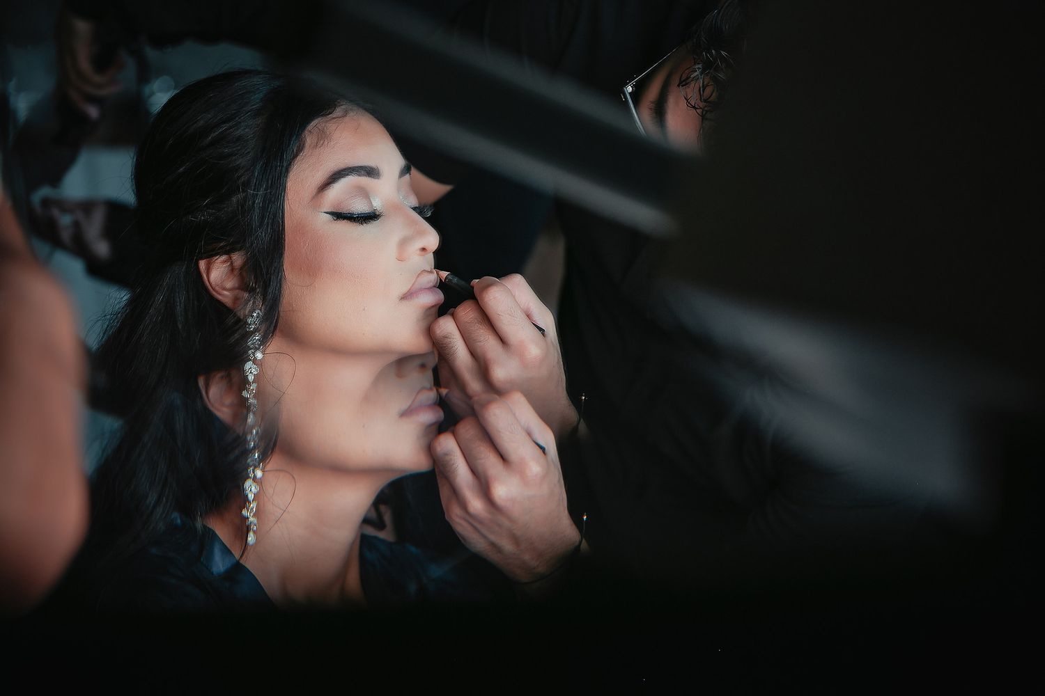 Professional makeup artist applies finishing touches to model's face in dramatic dark lighting.