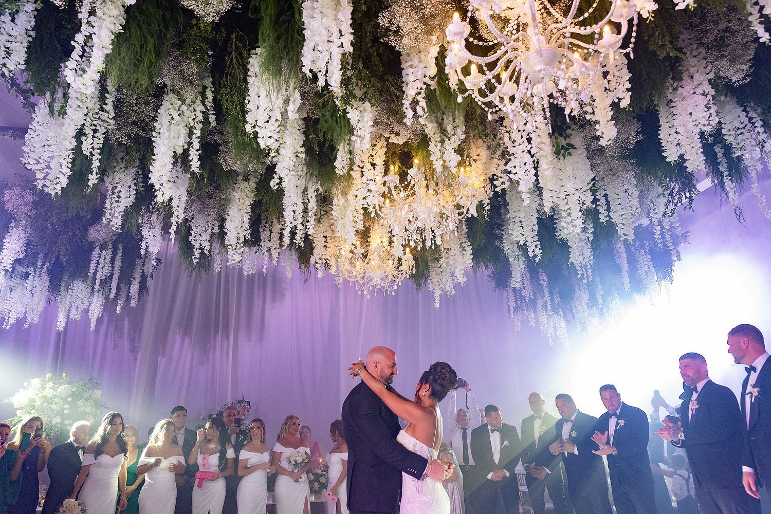 A couple shares their first dance under a stunning canopy of cascading white wisteria flowers and elegant chandeliers.