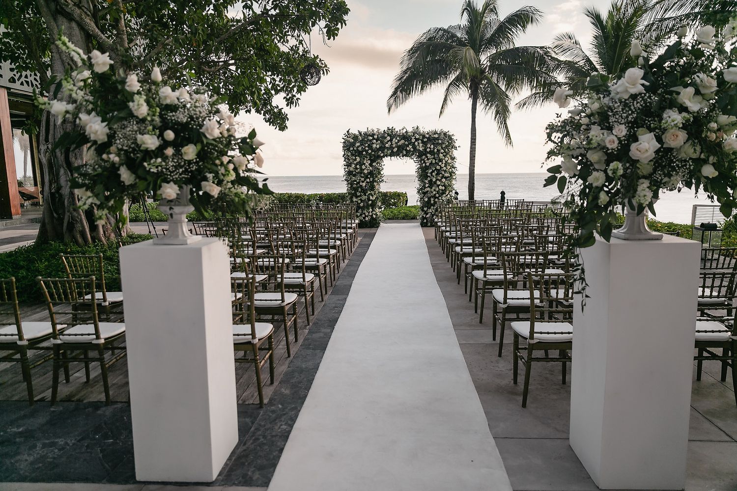 Outdoor wedding ceremony setup with white chairs arranged in rows along a palm tree-lined path.