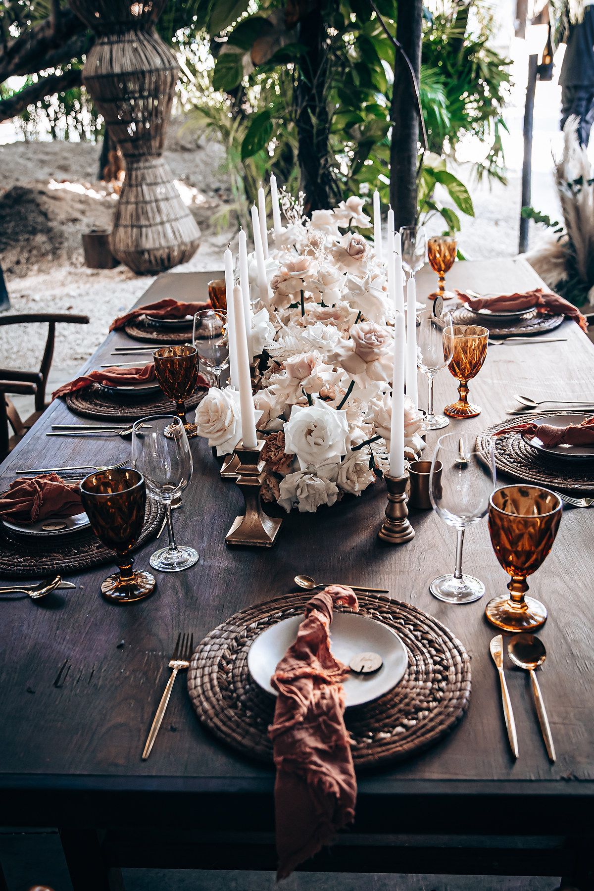 Elegant table setting with amber glassware, woven placemats, and white floral centerpieces on a dark wooden table.