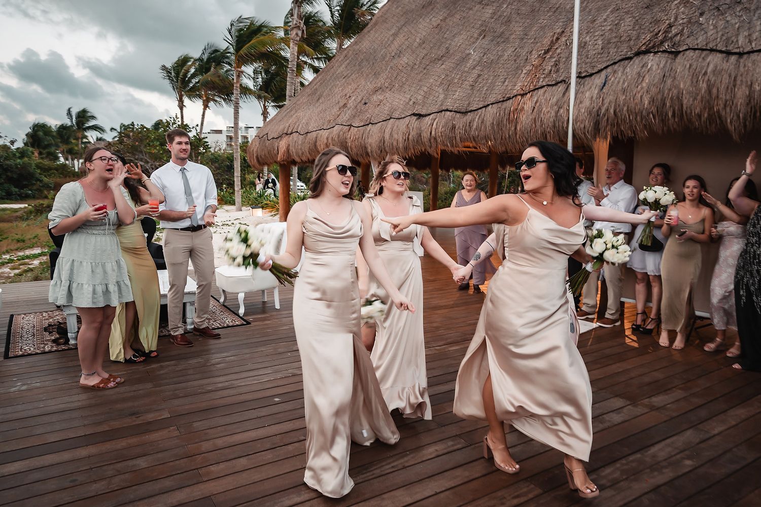Wedding guests dancing and celebrating on an outdoor wooden deck under a thatched roof.