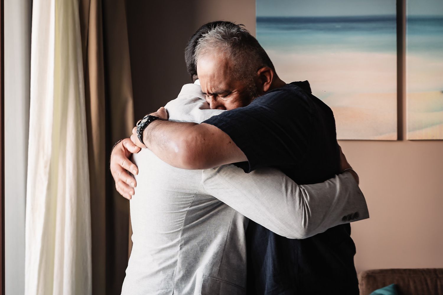 Two men sharing an emotional hug in a warmly lit room with a coastal painting in the background.