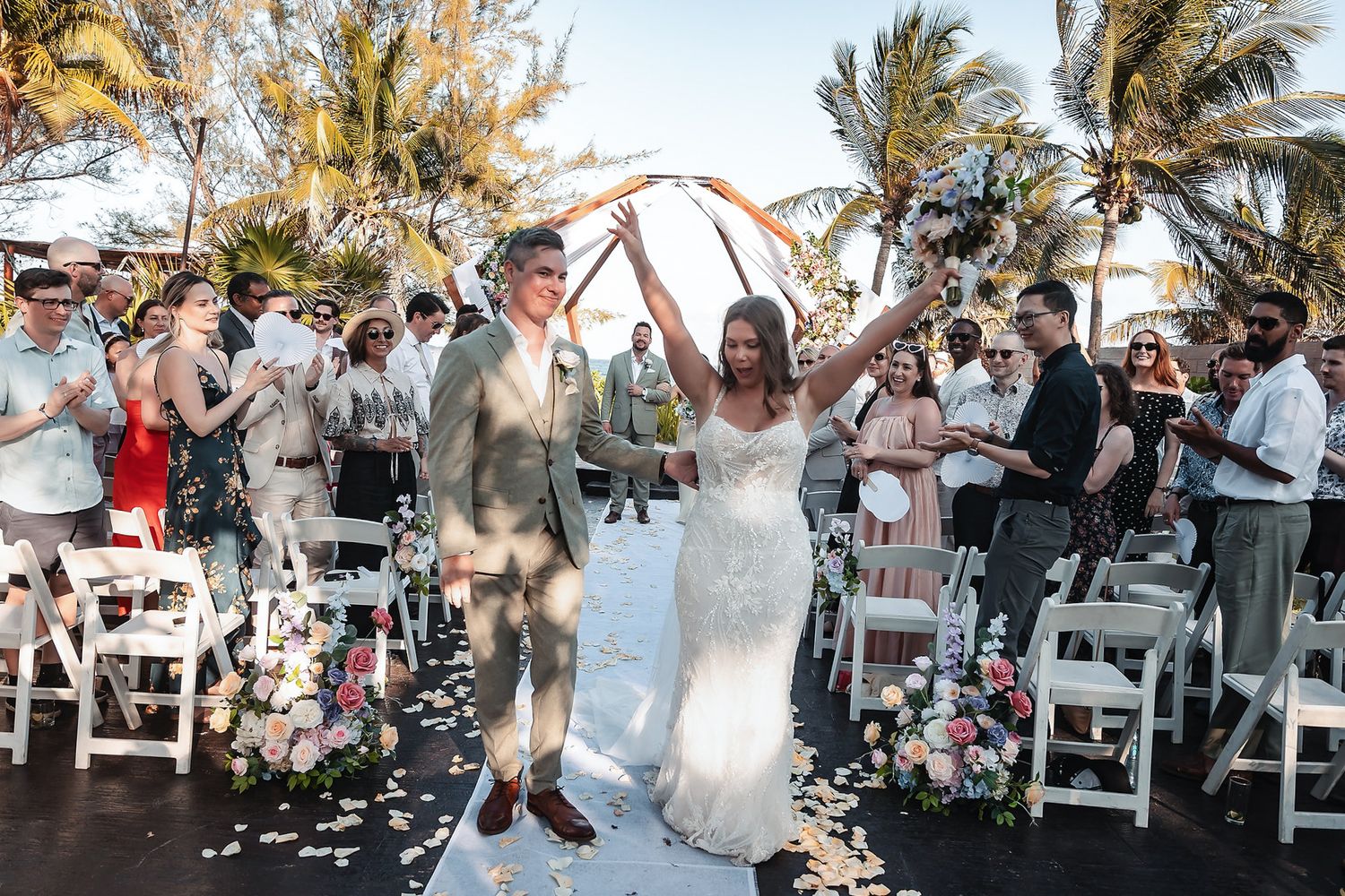 Joyful bride raises arms as couple exits wedding ceremony on tropical beach with palm trees.