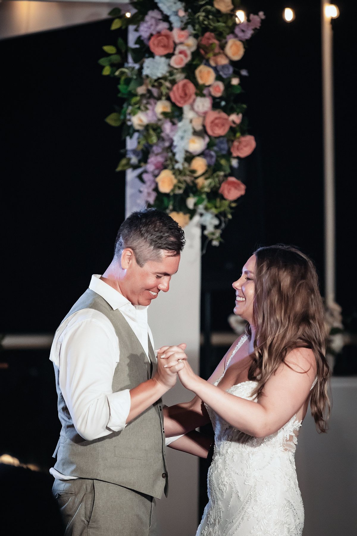 Bride and groom share a joyful first dance, laughing together beneath a floral arrangement at their wedding reception.