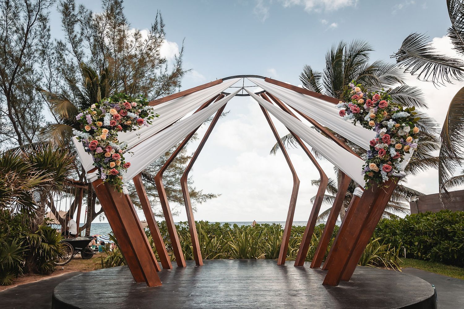 Elegant geometric wedding arch adorned with floral arrangements and white draping on a beachfront.