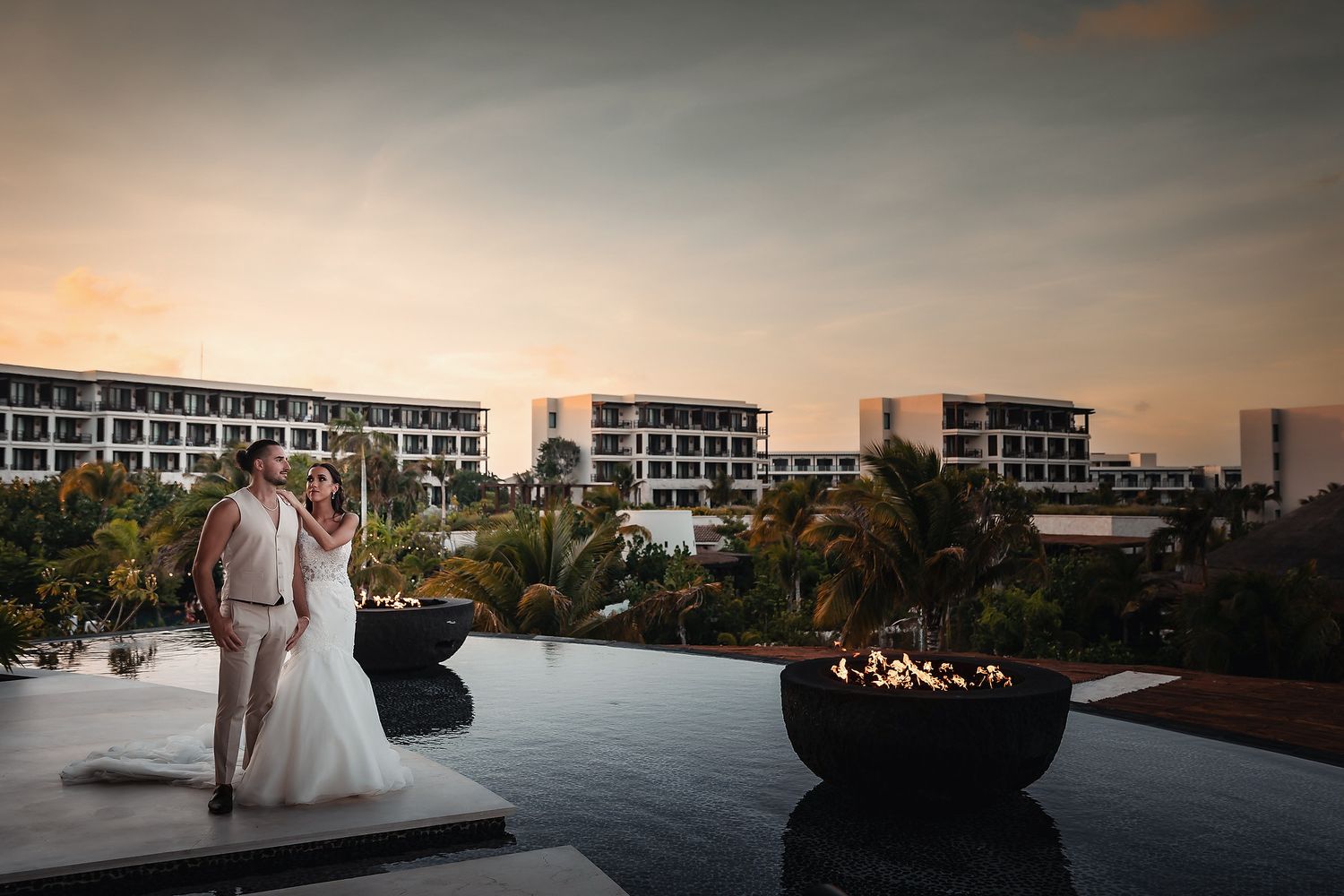 Bride and groom share a romantic sunset moment by a resort pool with fire bowls and tropical landscape backdrop.