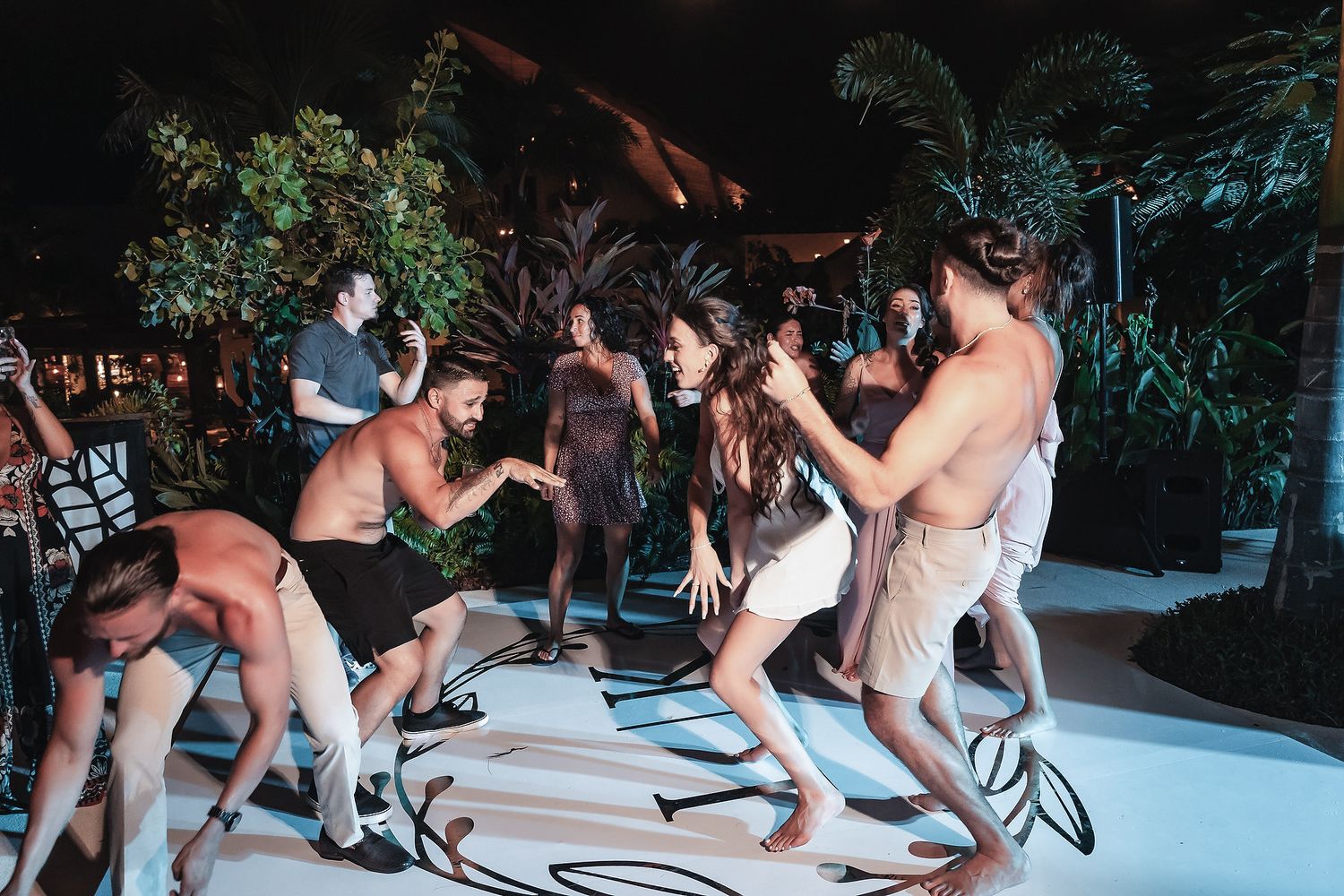 Group of people dancing energetically at an outdoor tropical night party with lush greenery backdrop.