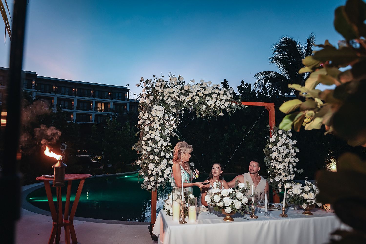 Elegant outdoor wedding reception by a pool at dusk with floral arch and sweetheart table.