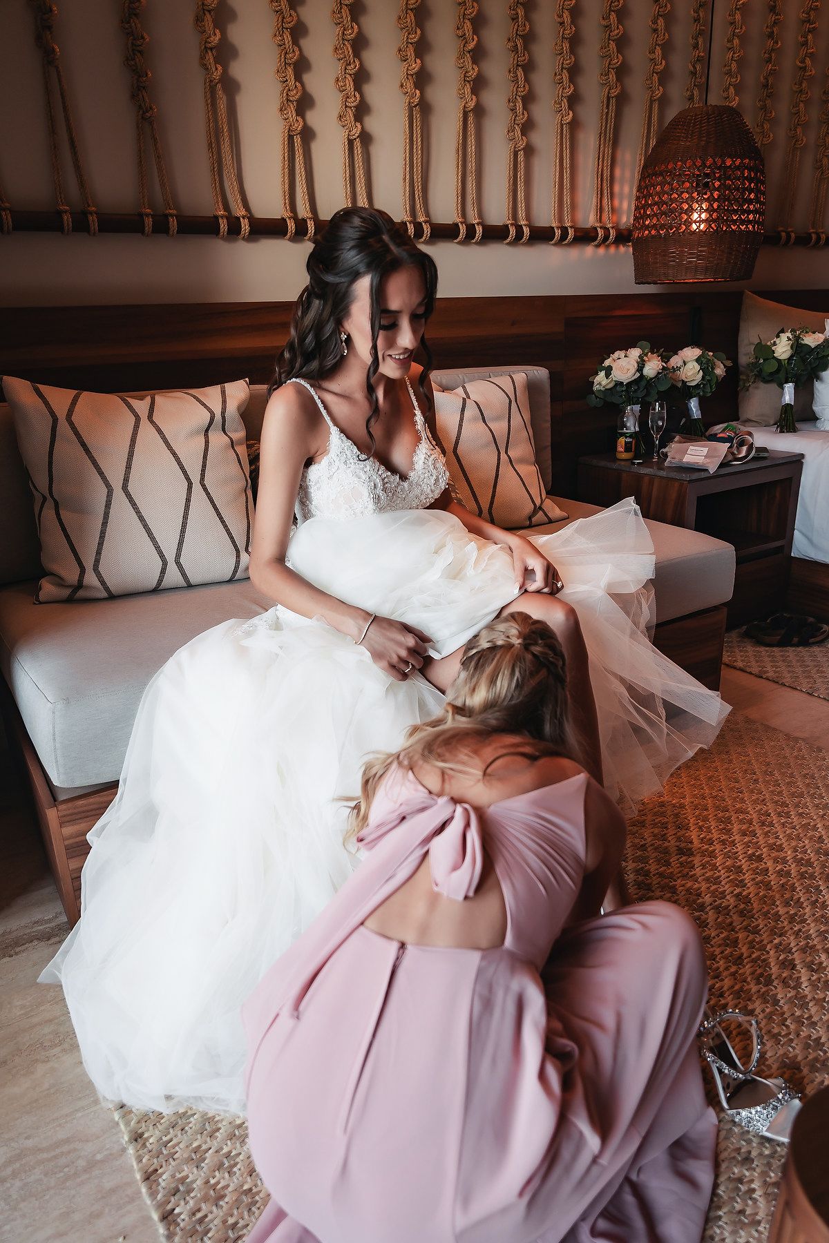 Bride in white lace gown sits smiling while bridesmaid in pink dress adjusts her dress in elegant hotel room.