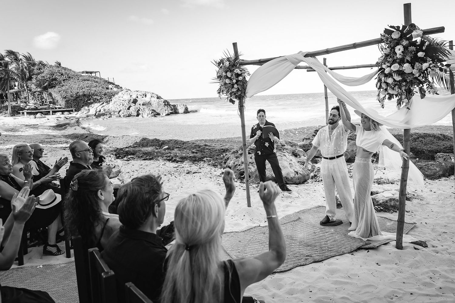 Black and white beach wedding ceremony with couple at floral arch, guests celebrating on sandy shore with ocean backdrop.