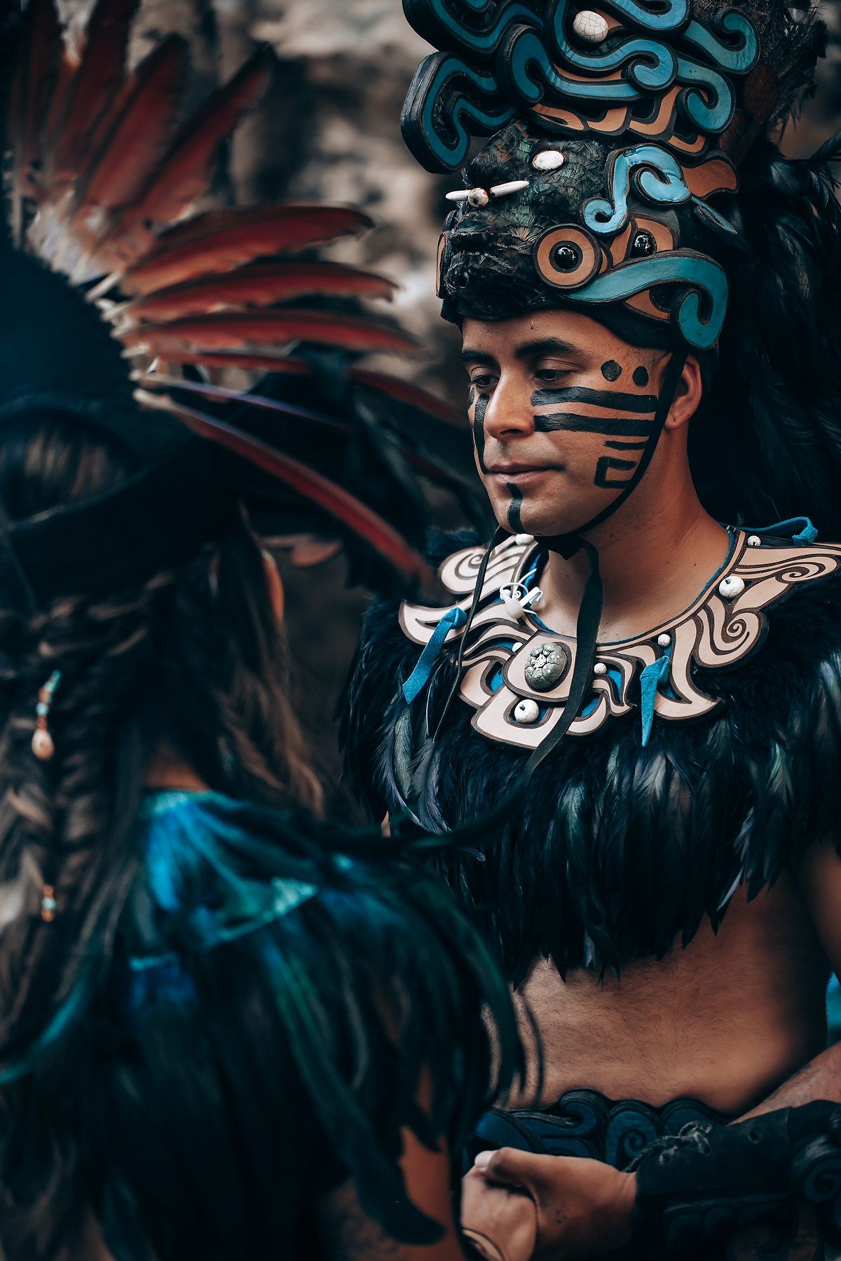 Male performer in detailed Mayan warrior costume with black feathers, face paint, and ornate turquoise headdress.