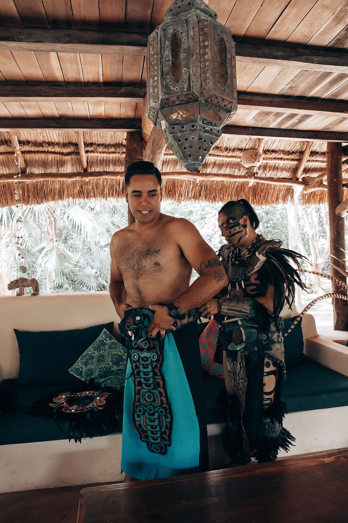 Shirtless man being dressed in traditional Mayan ceremonial attire by a performer in elaborate indigenous costume.