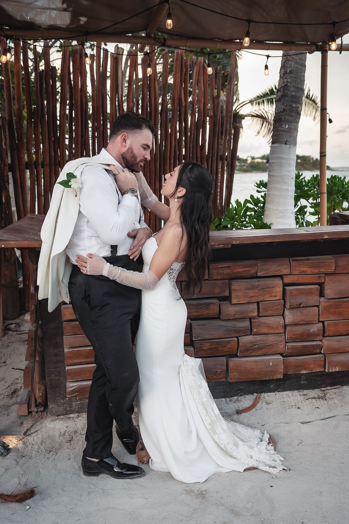 Bride and groom share a romantic moment by a rustic beach bar, with palm trees and string lights in the background.