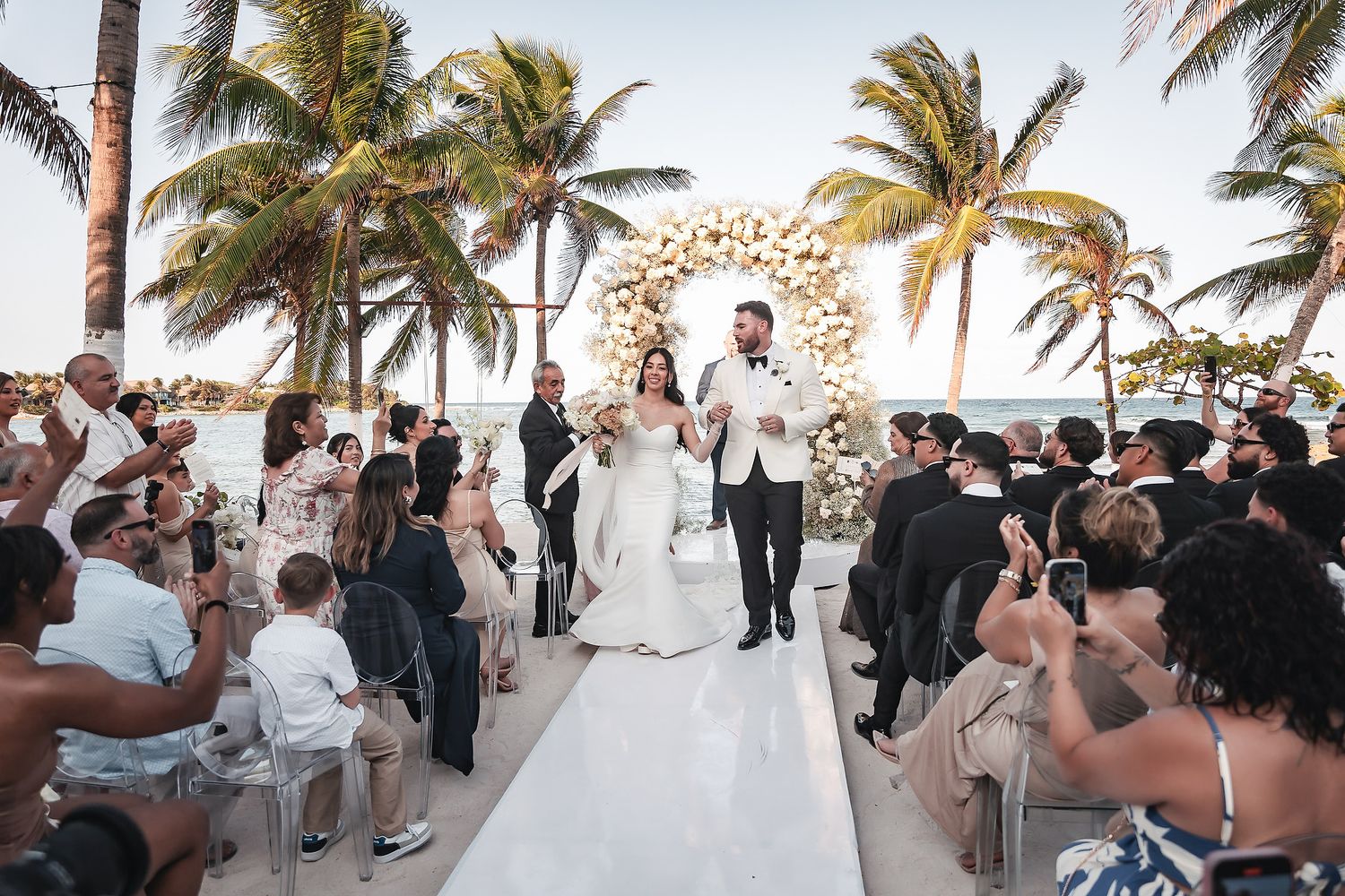 Newlyweds walk down white aisle at outdoor beach wedding ceremony surrounded by guests and palm trees.
