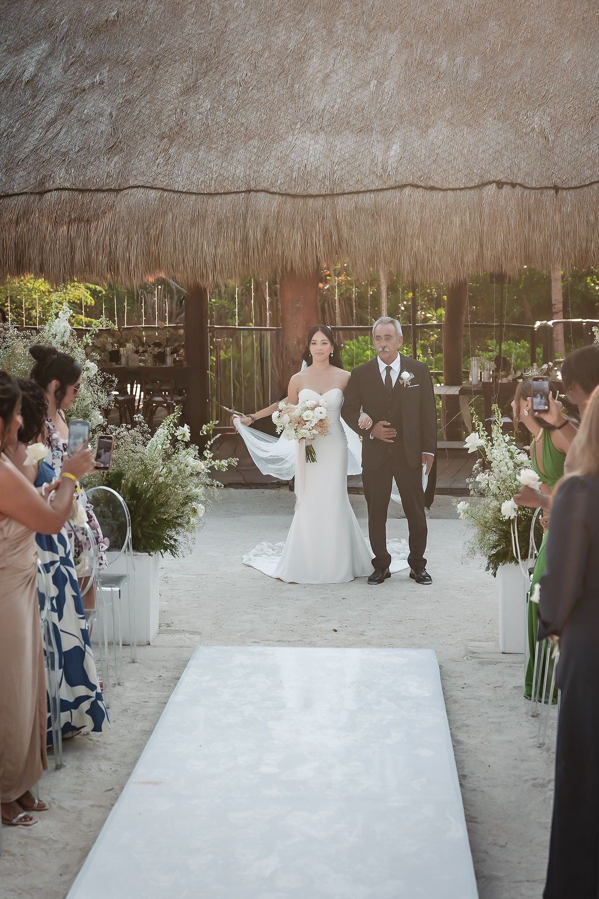 Bride walks down sandy aisle with escort under thatched roof at elegant beach wedding ceremony.