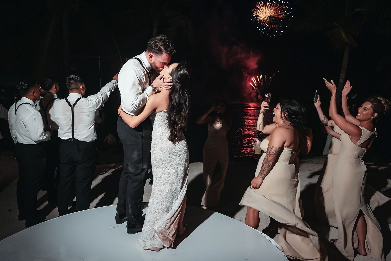 Bride and groom share a romantic kiss on the dance floor surrounded by guests and colorful fireworks at their wedding reception.