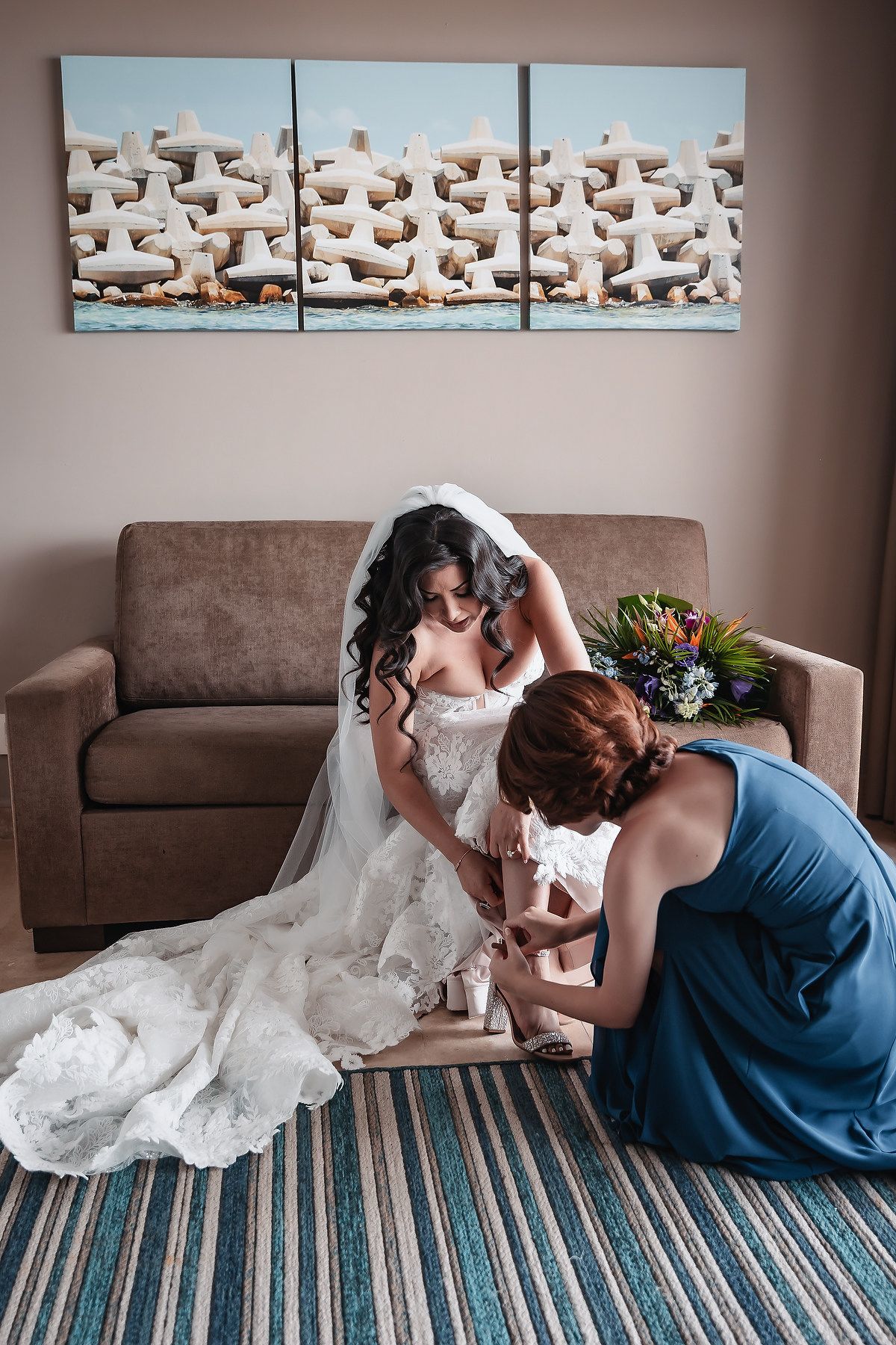 Bridesmaid in blue dress helps bride with shoes while seated on striped rug in hotel room before wedding ceremony.