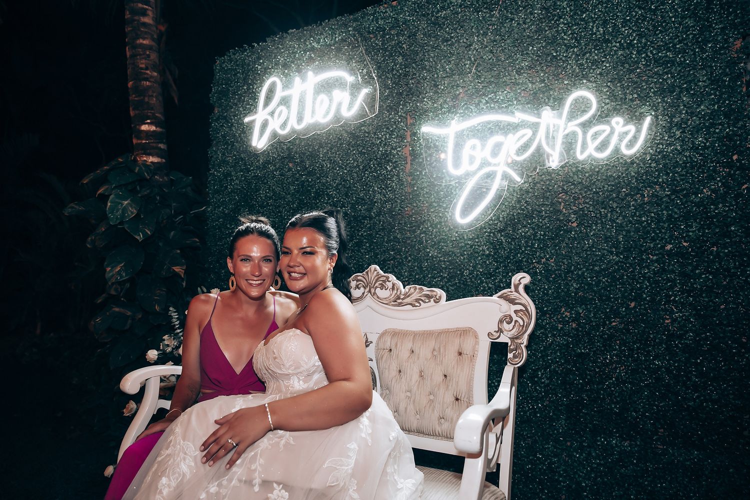 Two women share a joyful moment on an elegant white chair against a backdrop with neon lighting.