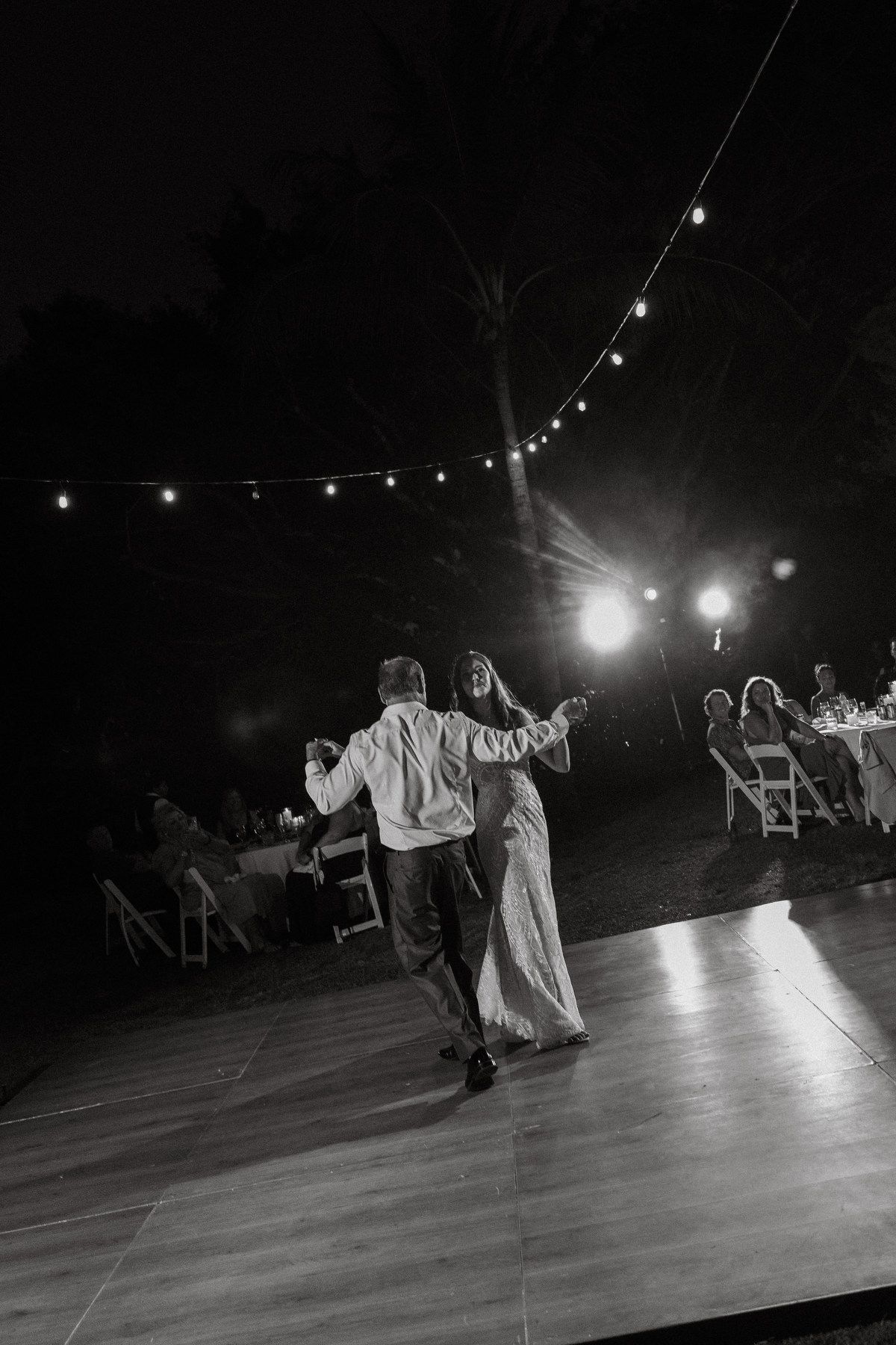 Bride and groom share elegant first dance under string lights on outdoor dance floor in black and white.