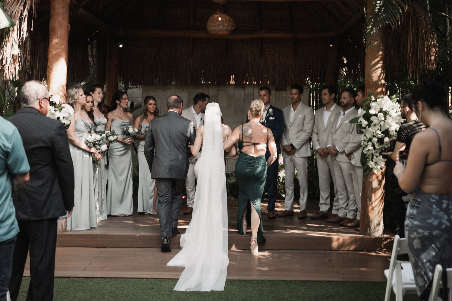 Bride walking down the aisle in a tropical palapa wedding venue with guests and wedding party standing on both sides.