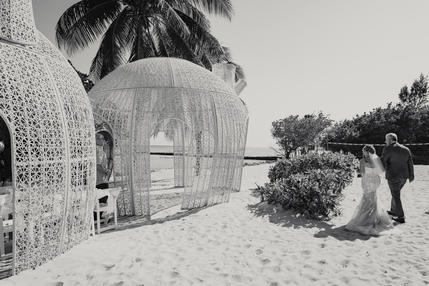 Black and white photo of bride and groom walking toward ornate beach wedding gazebo with palm trees.