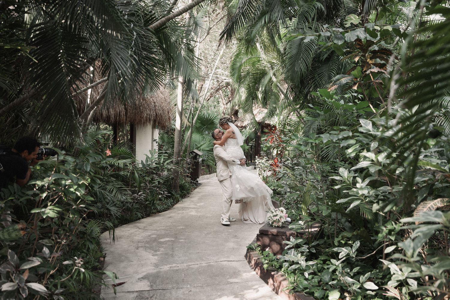 Newlywed couple embraces and twirls on a lush tropical garden pathway surrounded by palm trees.