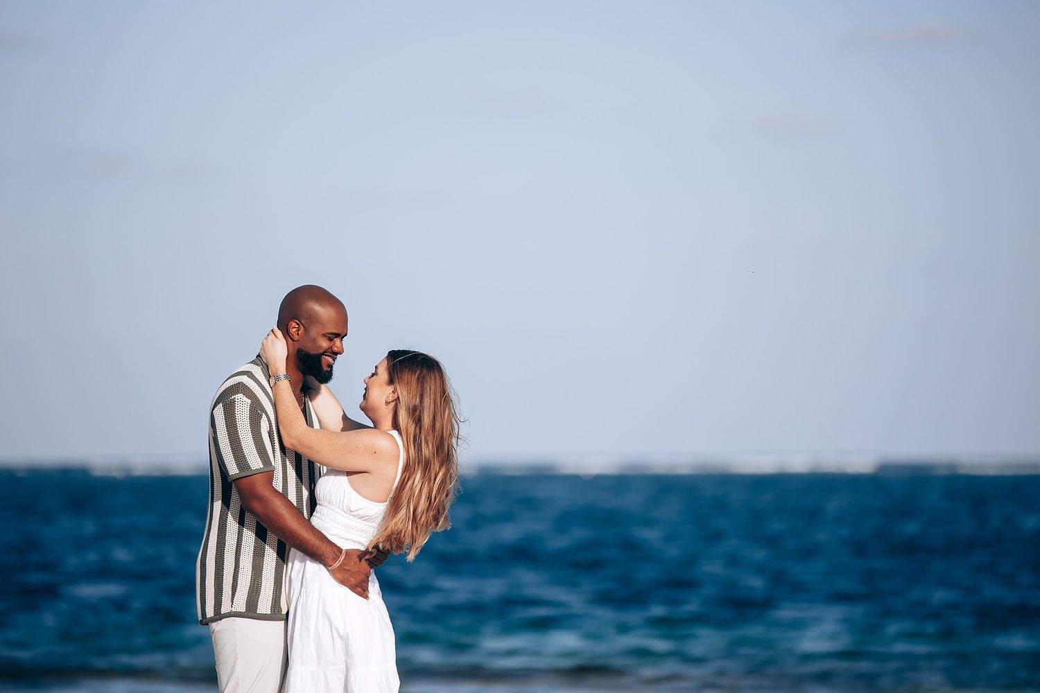 Interracial couple embracing romantically on a tropical beach with turquoise ocean in the background.