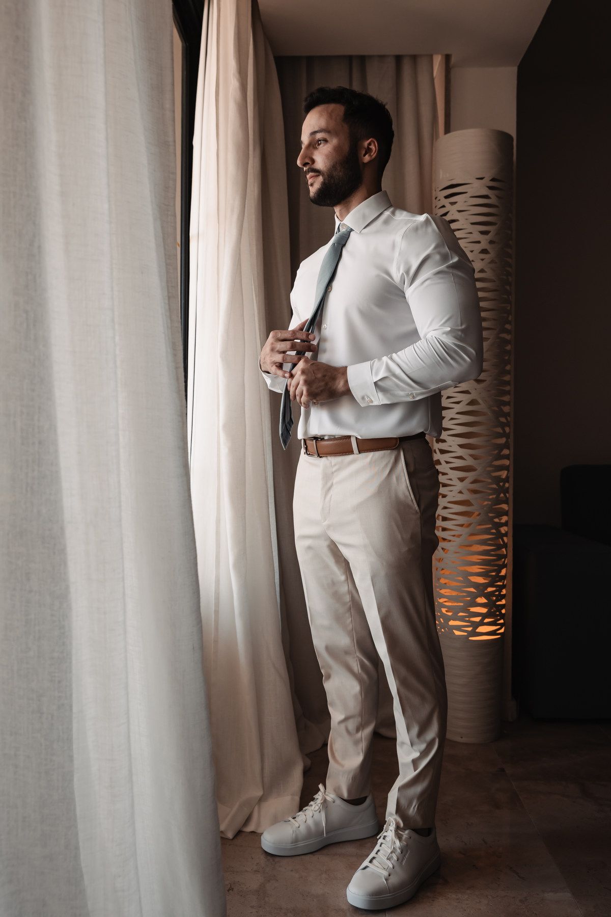 Groom in white shirt and beige pants adjusting his tie while standing by a window in a hotel room.