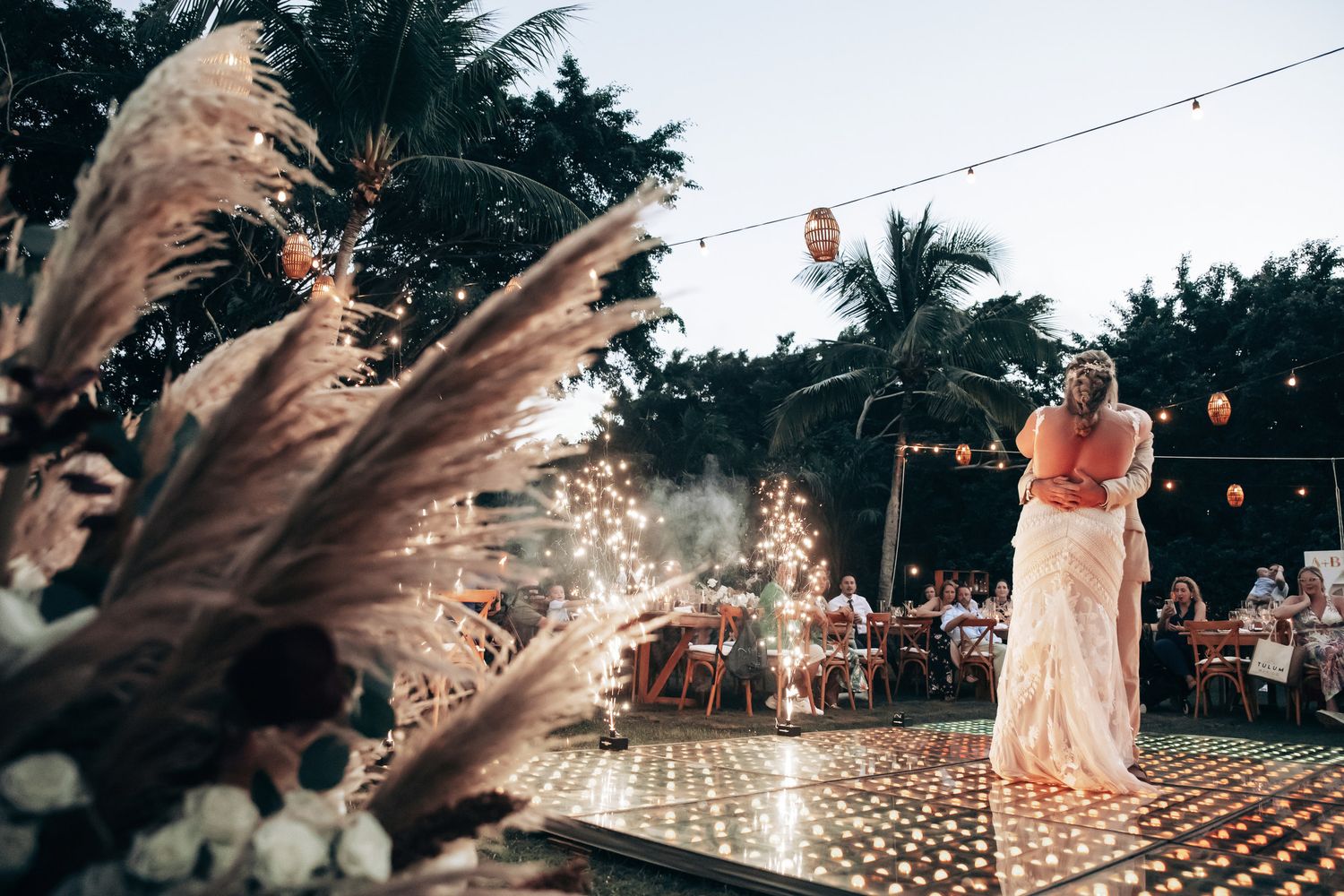 Bride stands on a glittering dance floor surrounded by pampas grass and string lights at a tropical evening wedding reception.