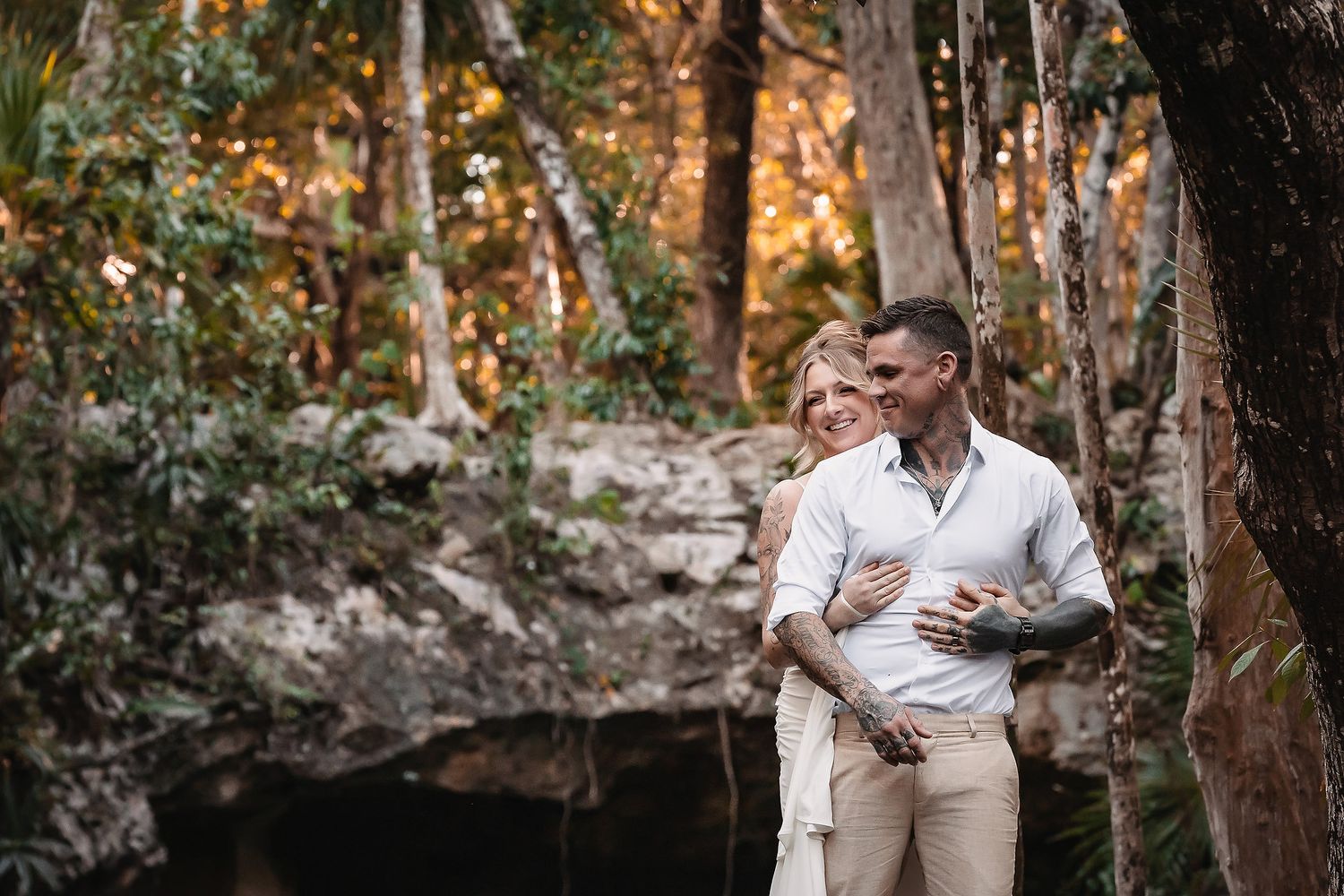Couple embracing in a lush forest setting, surrounded by trees and rocks, sharing a romantic outdoor moment.