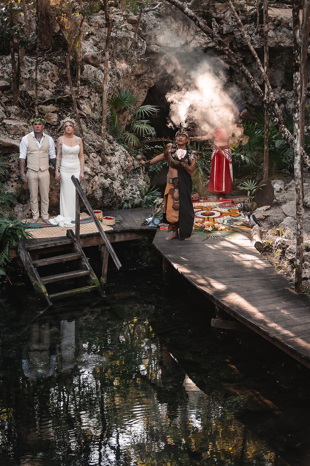 Mayan ceremony on a jungle cenote dock with couple, officiant burning incense, and red-robed figure amid lush greenery.