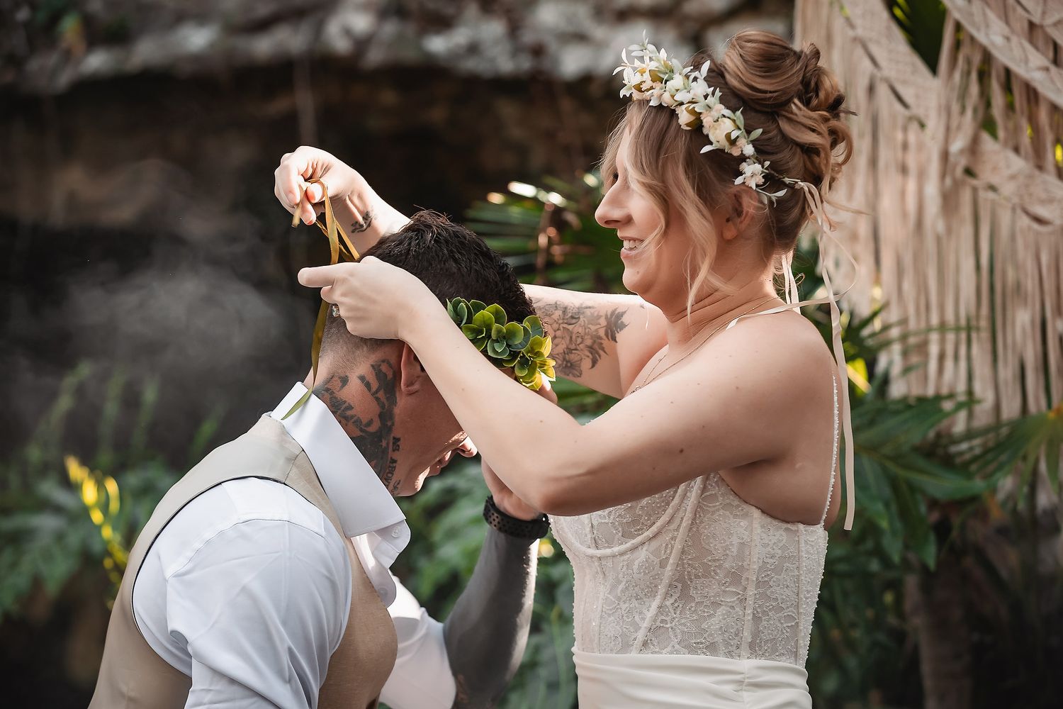 Bride placing floral crown on groom during outdoor wedding ceremony surrounded by tropical greenery.