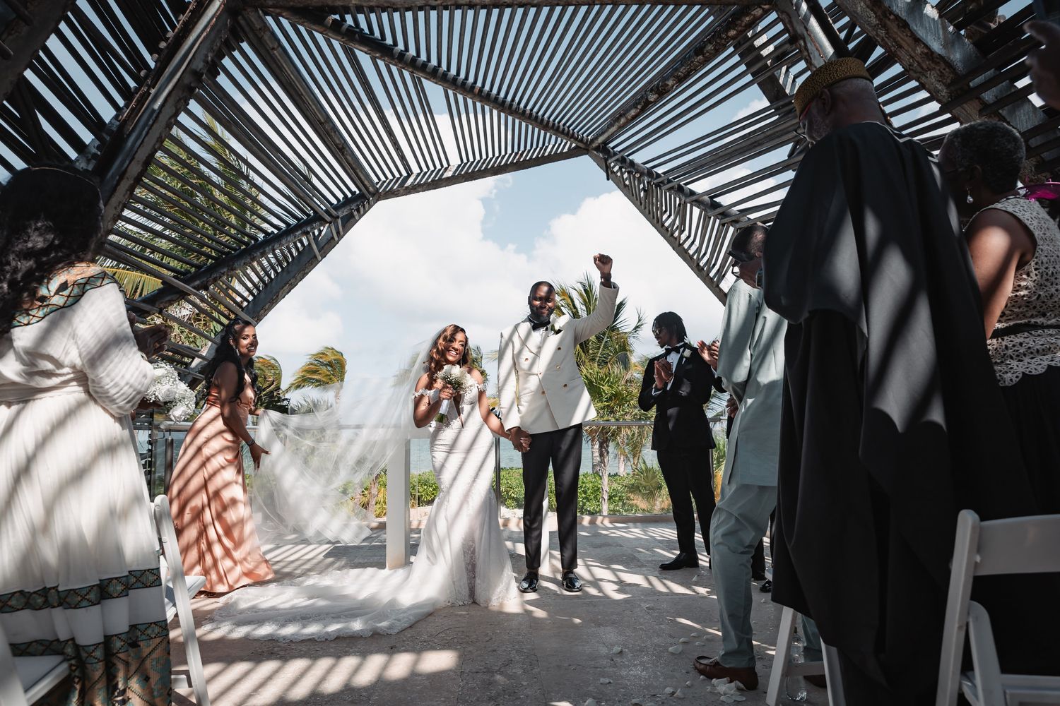 Newlyweds celebrate joyfully as they exit their outdoor wedding ceremony under a wooden canopy.