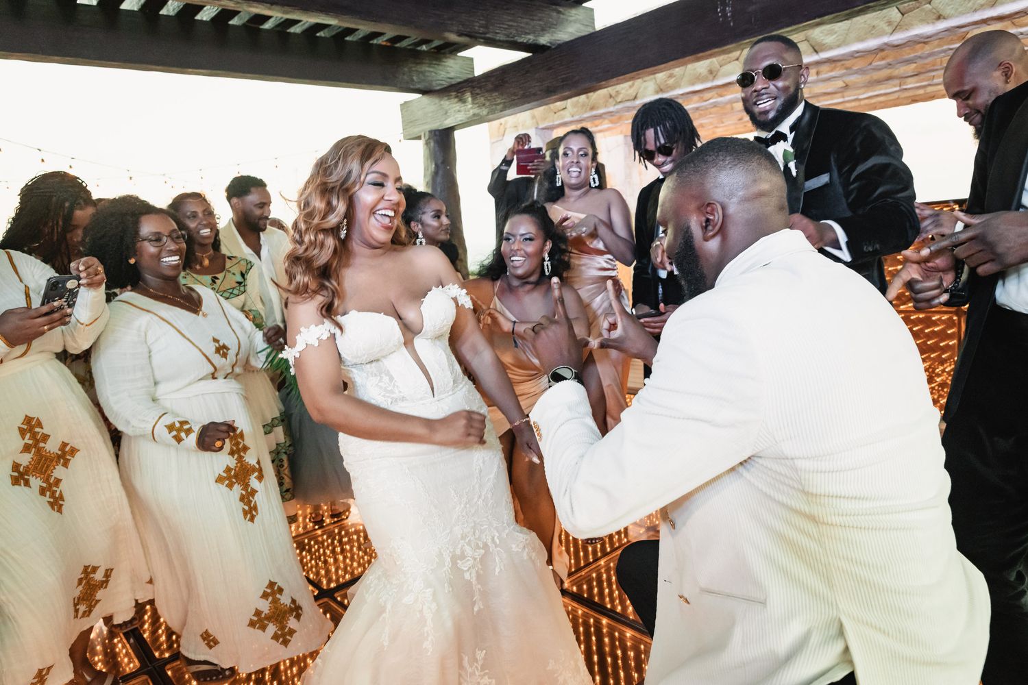 Groom kneels before smiling bride surrounded by bridesmaids in gold kente-trimmed dresses.