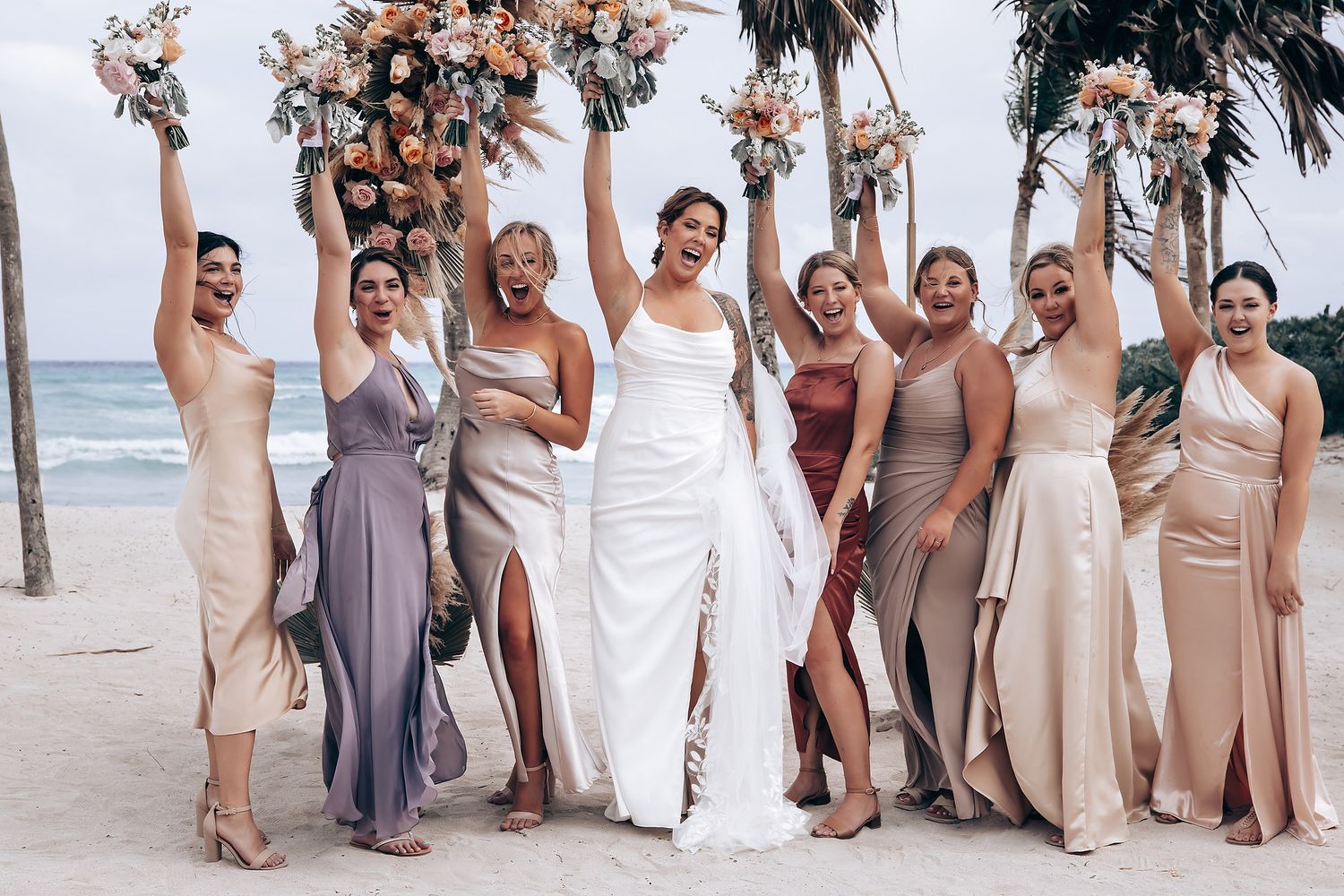 Bridal party in beige and lavender dresses stands together on sandy beach with palm trees in background.