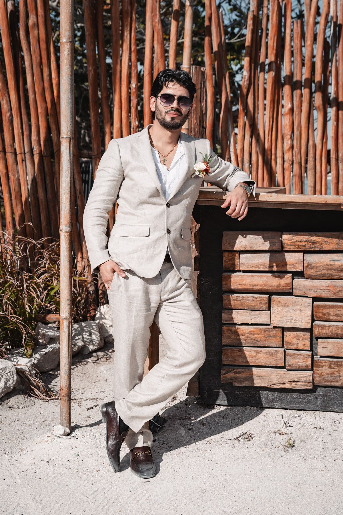 Stylish groom in beige linen suit with sunglasses leaning against wooden bar on sandy beach.