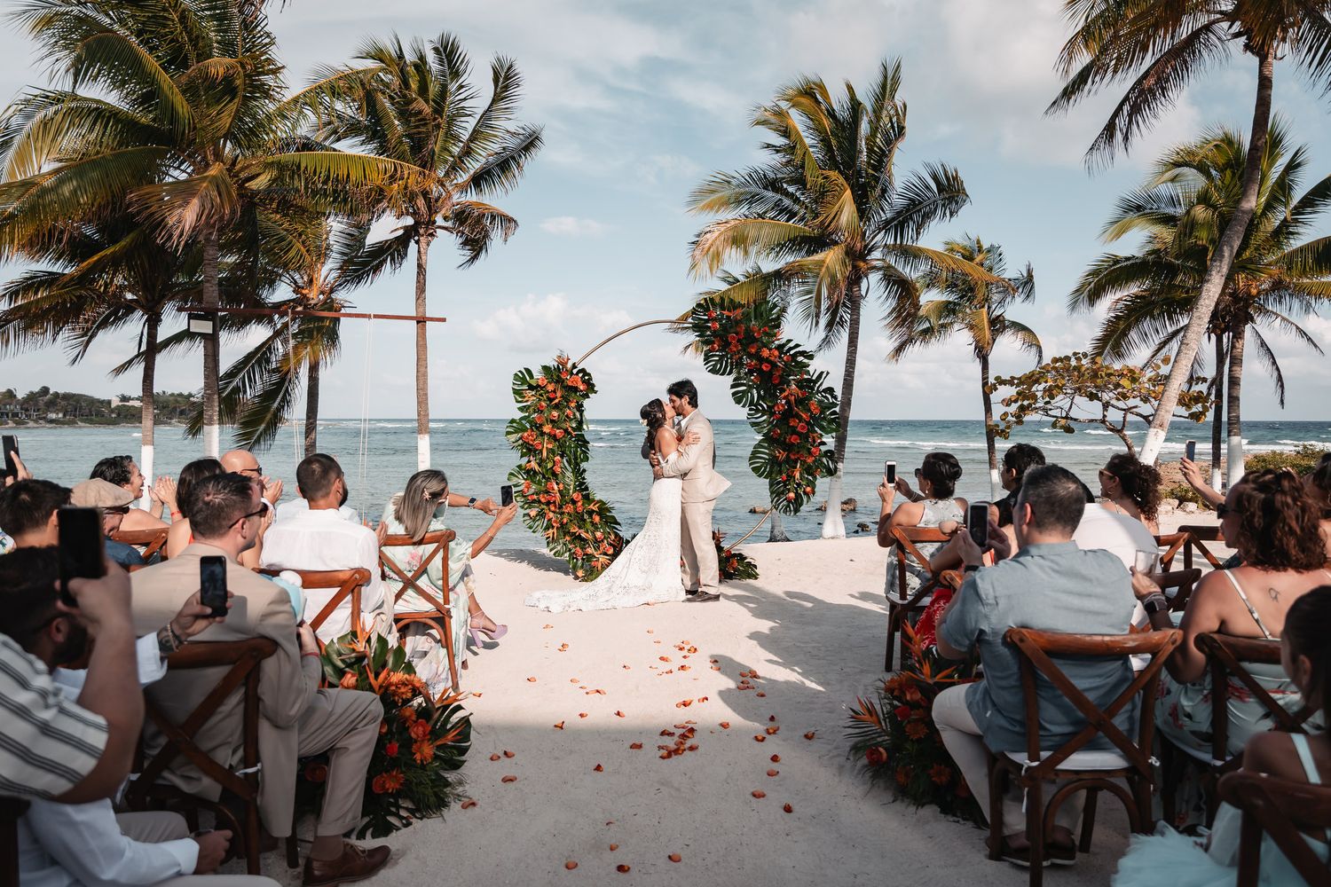 Couple kissing at beachfront wedding ceremony with palm trees and floral arch.