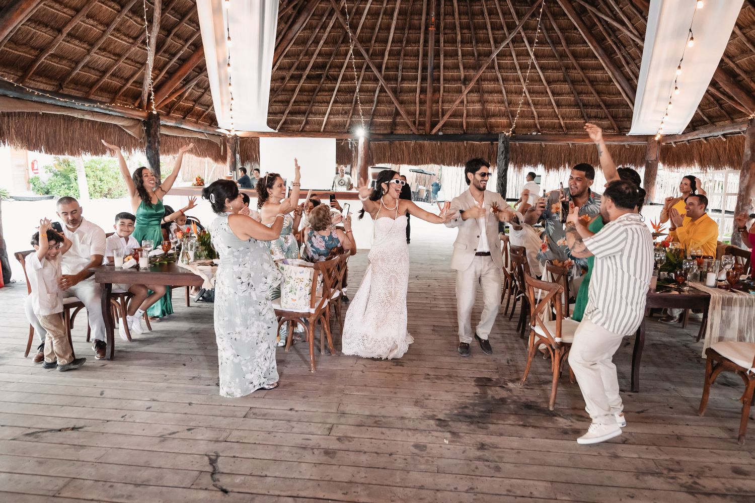 Bride and guests dancing joyfully under a thatched roof at beach reception.