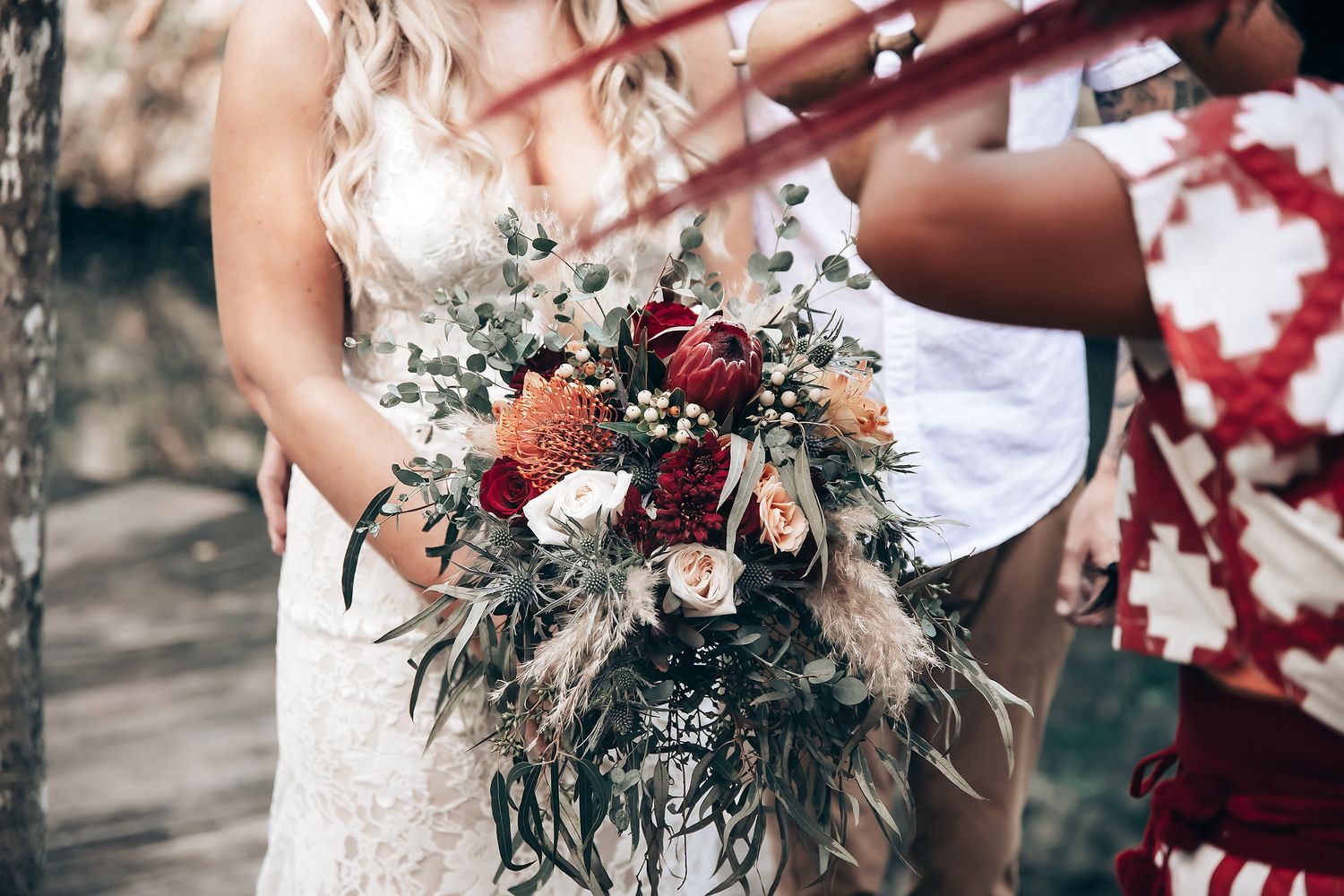 A wedding party stands together outdoors holding burgundy and white floral bouquets in a natural setting.