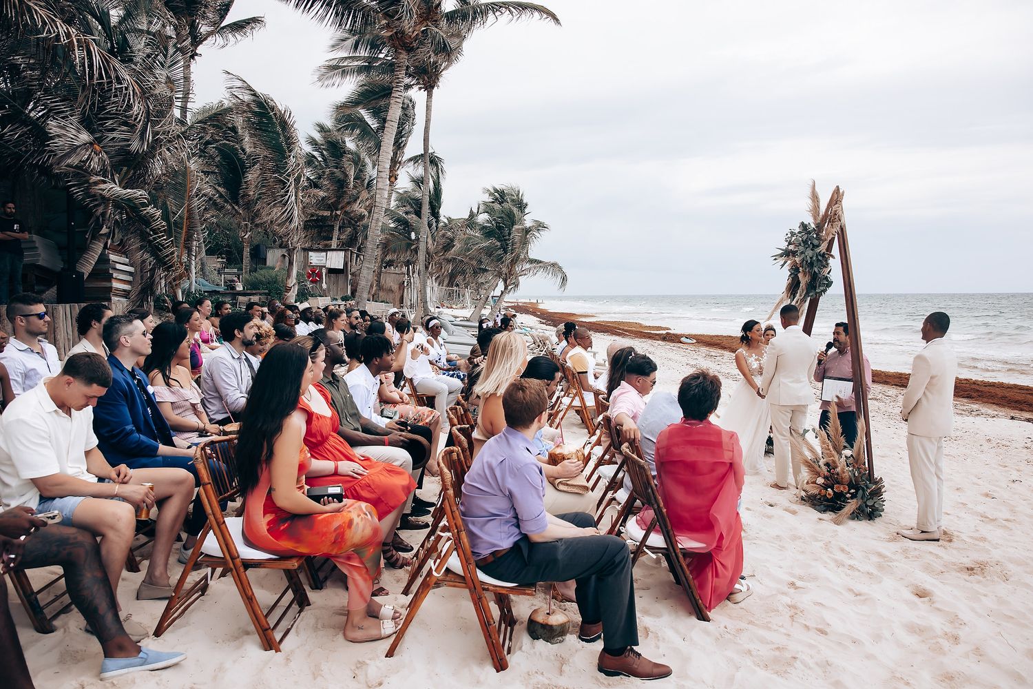 Beachside wedding ceremony with guests seated under palm trees.