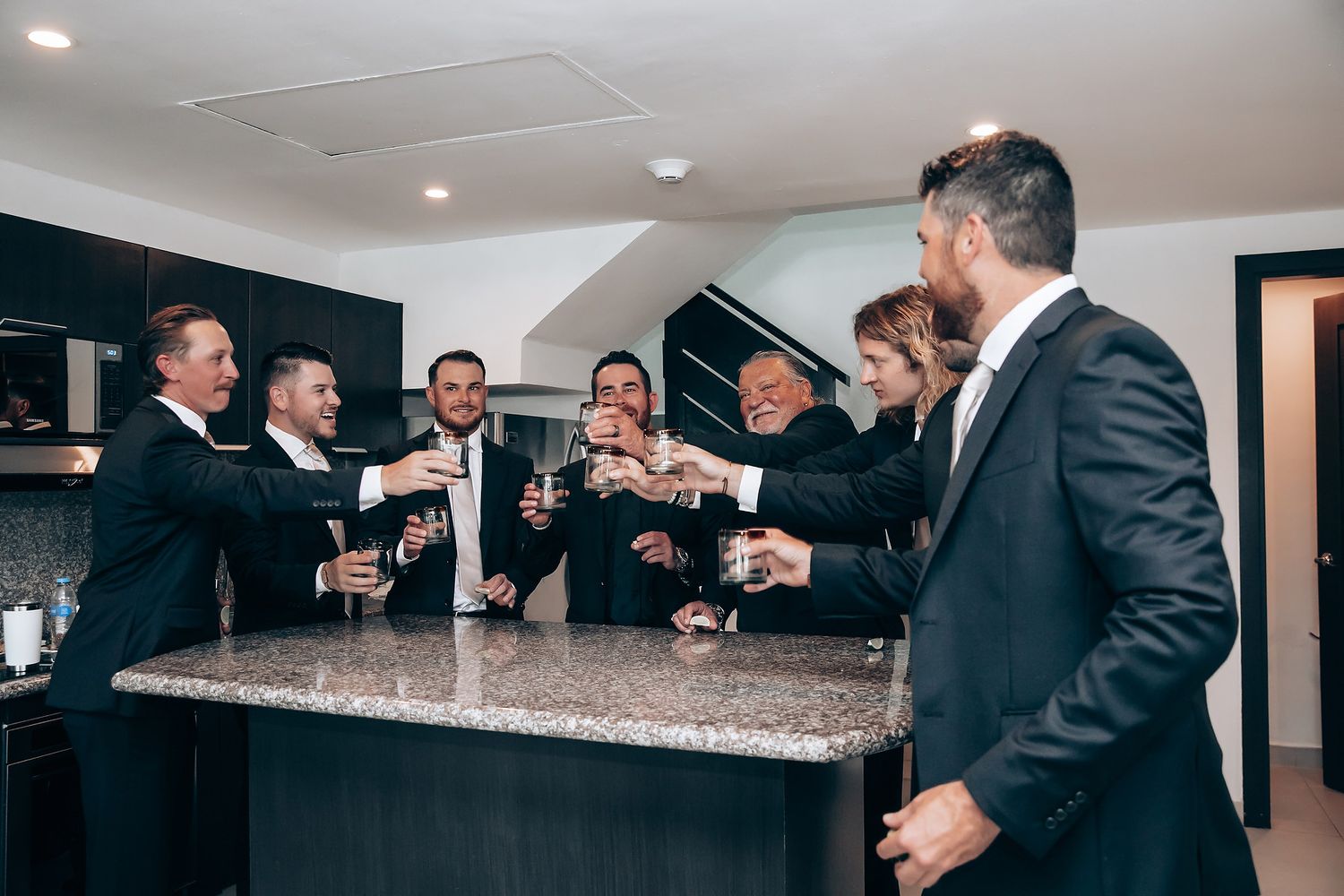 Wedding party members gathered around a bar counter sharing drinks and laughter.