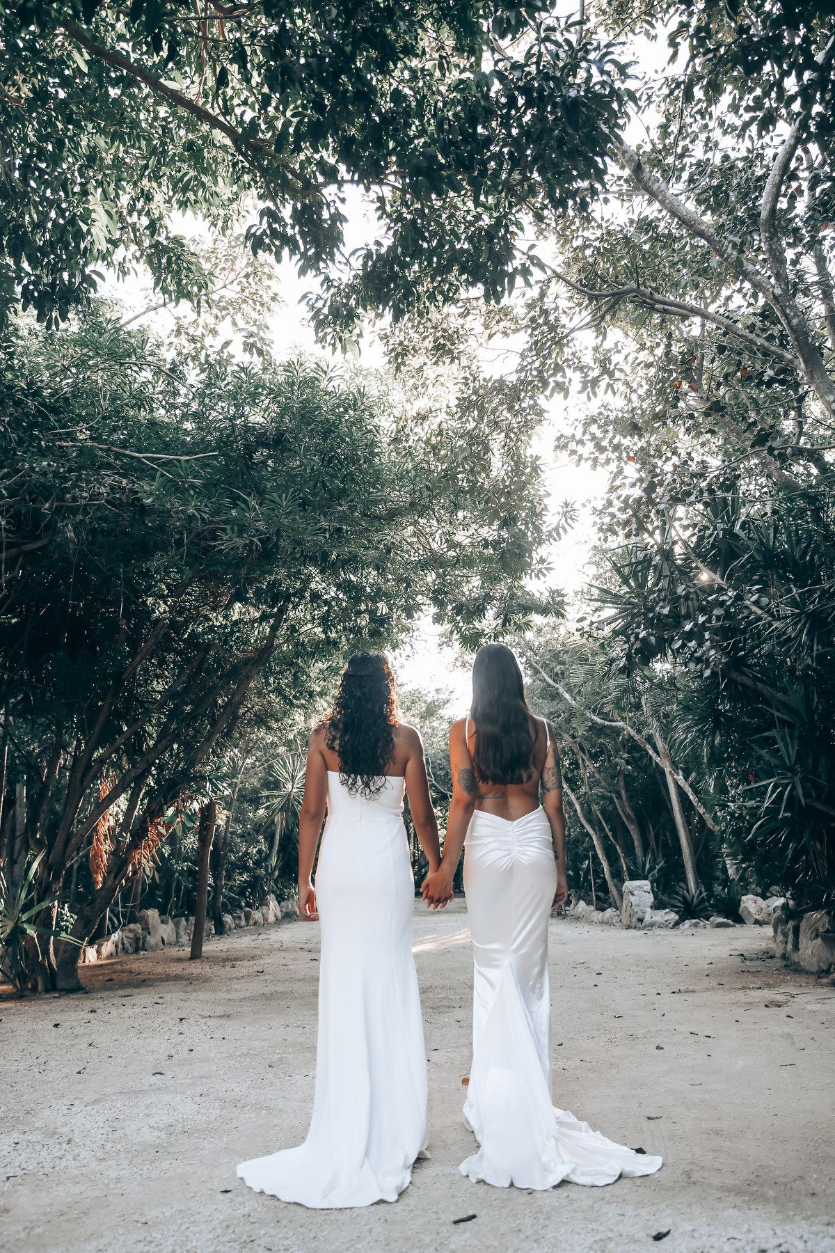 A couple in matching white dresses stands together on a tree-lined beach path.