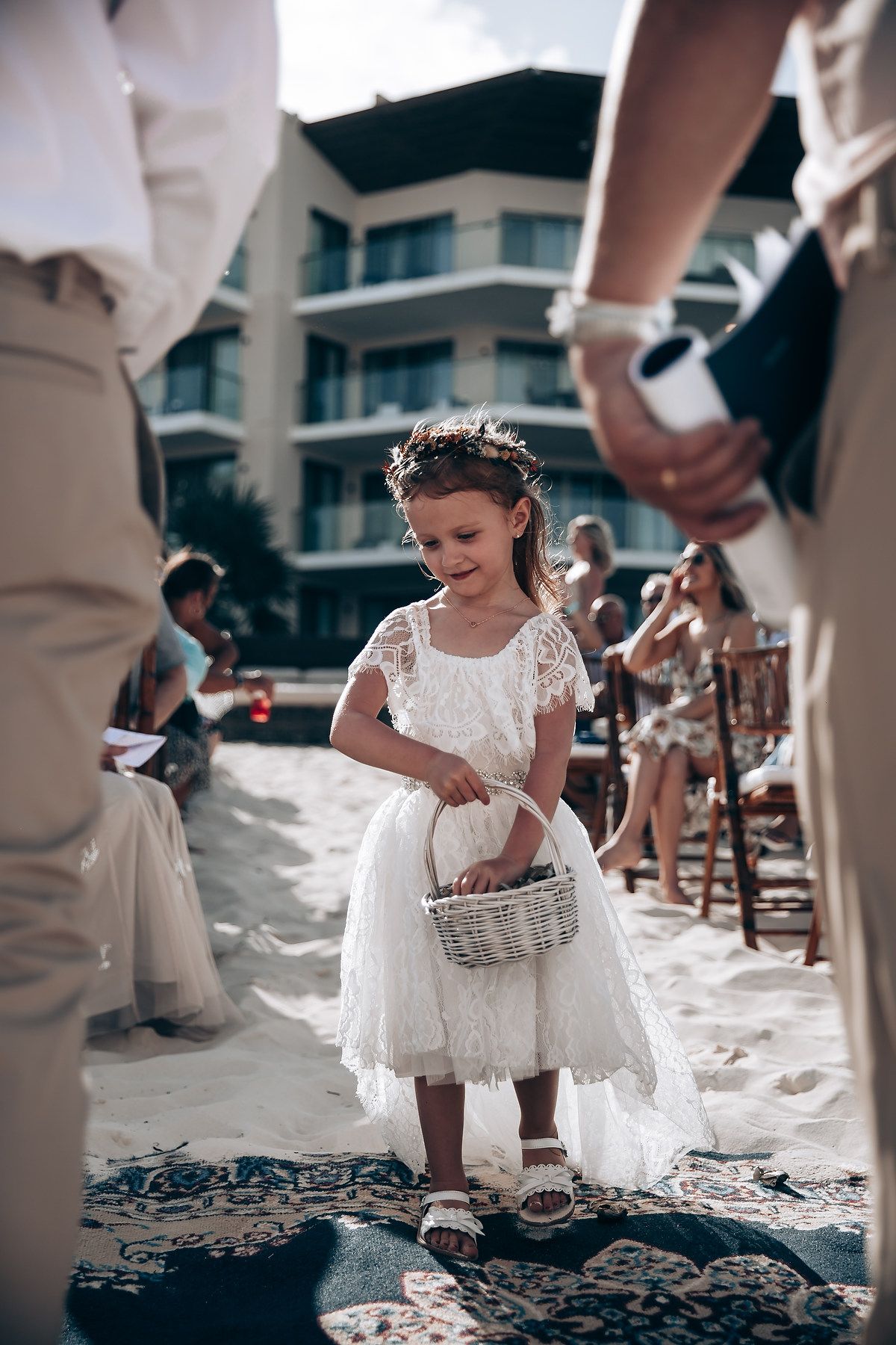 A young flower girl in a white lace dress scatters petals on a beach during a wedding ceremony.