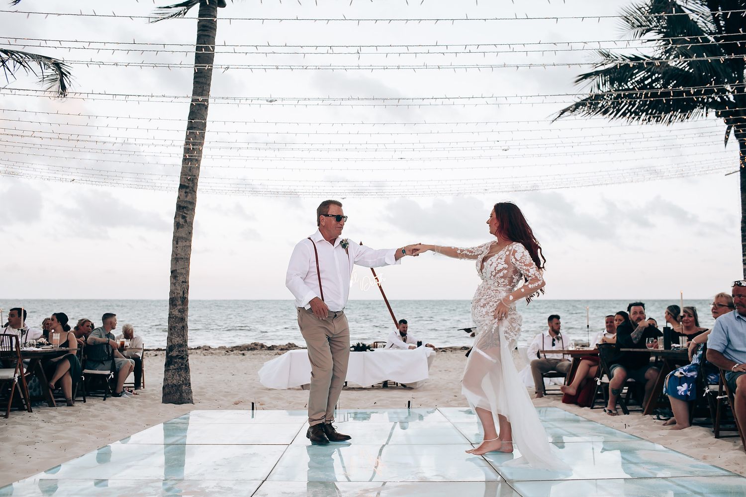 Wedding couple dancing on white platform at beachside reception with palm trees and guests watching.