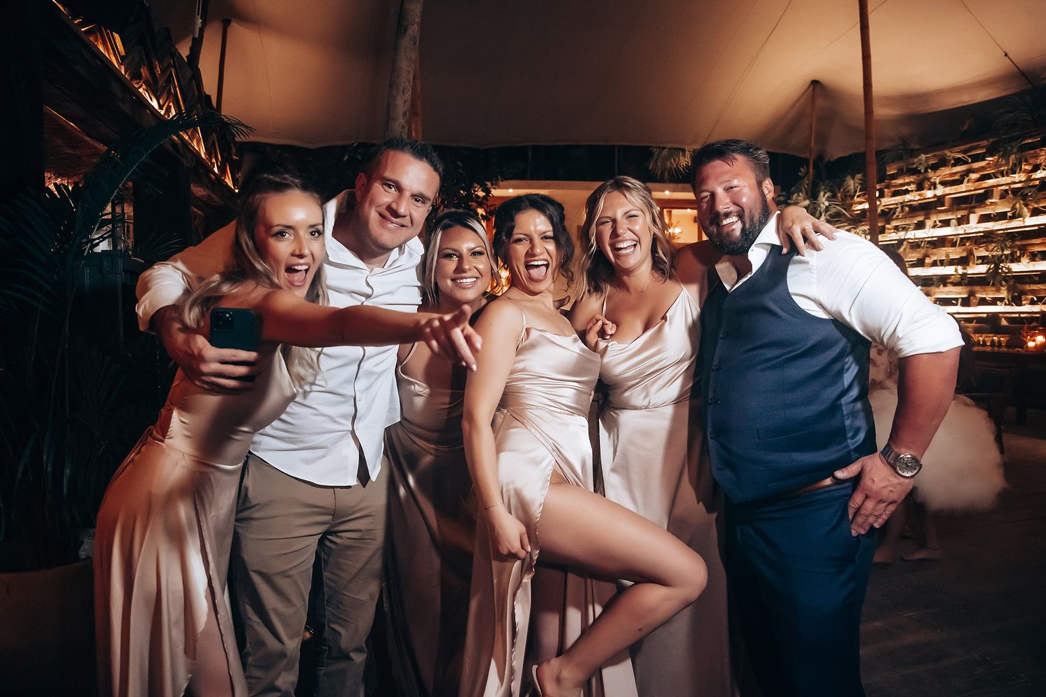 Wedding party celebrates on dance floor under tent with string lights and festive atmosphere.