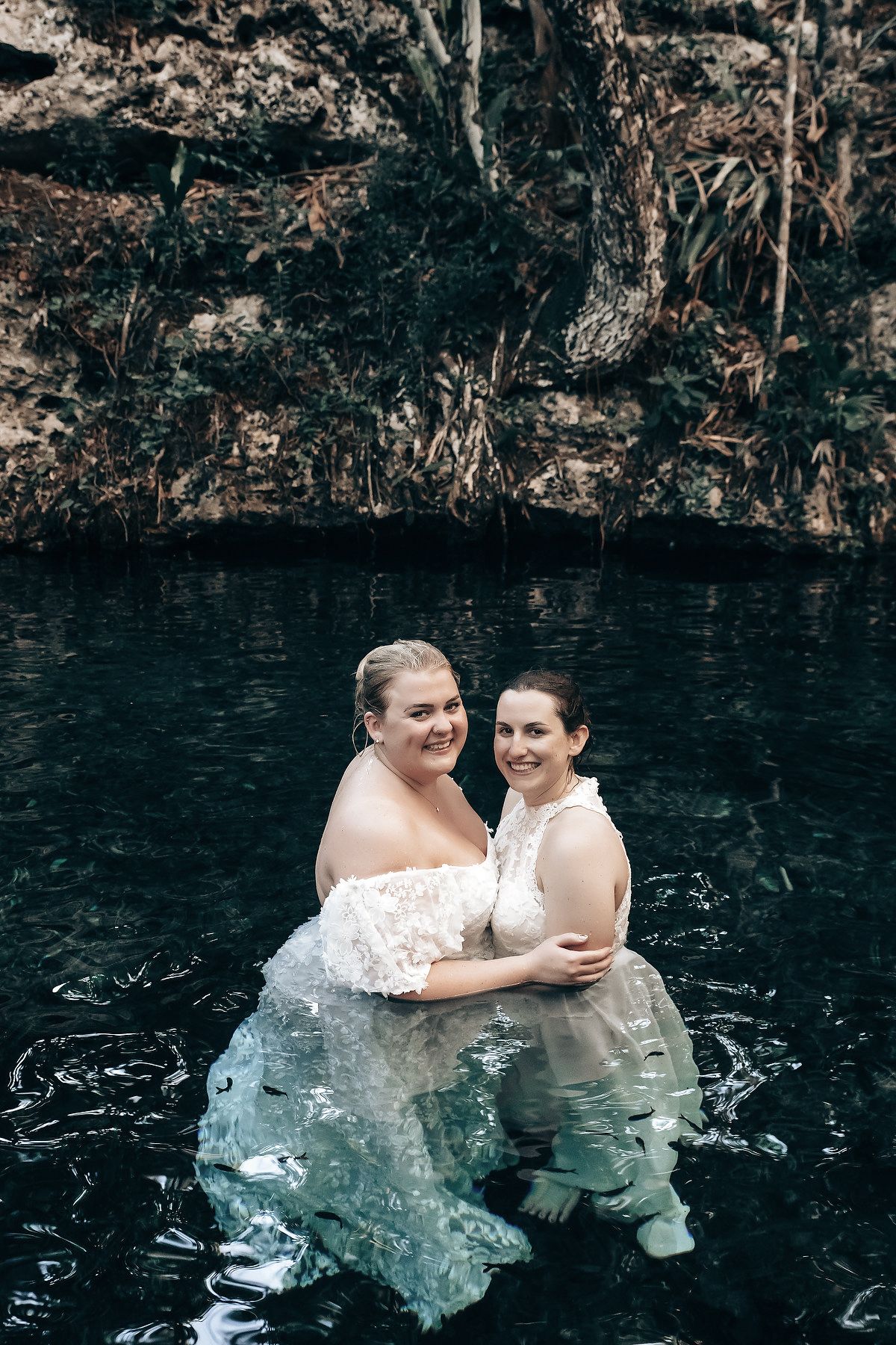 Two women in wedding dresses standing in a body of water surrounded by a natural landscape.