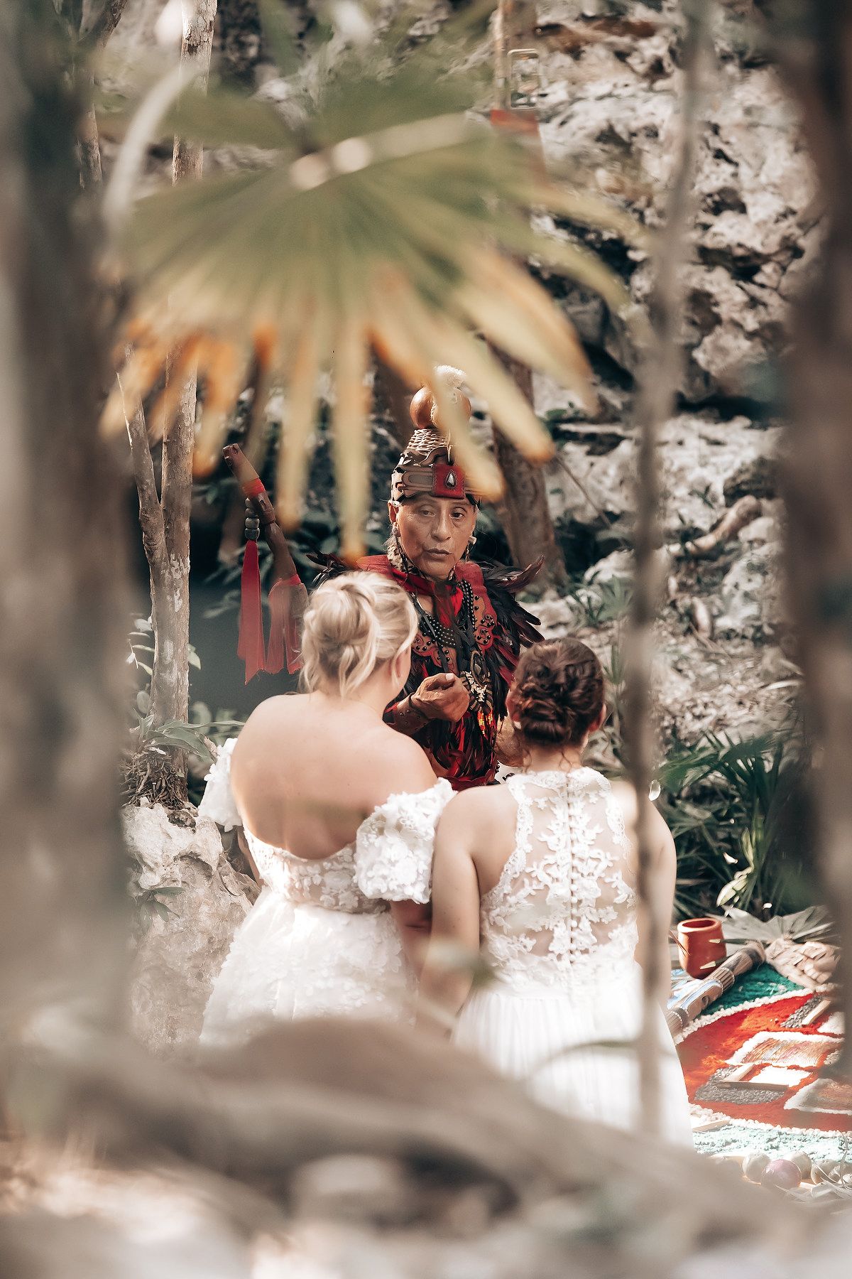 The image shows a bride and groom standing in a forest setting, with a person wearing a colorful, ornate costume in the background.