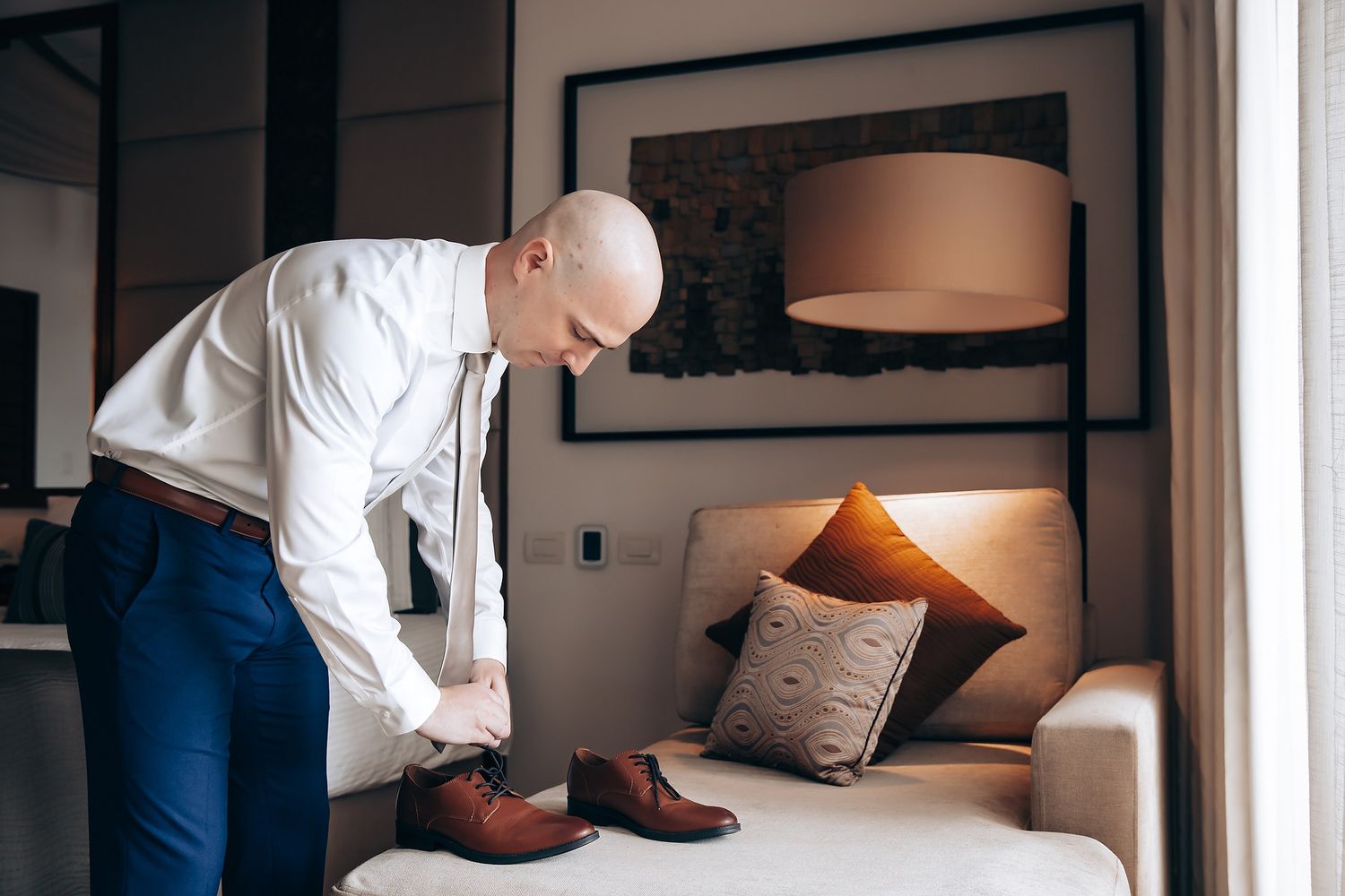 A man in a white shirt bends down to put on a pair of brown shoes.